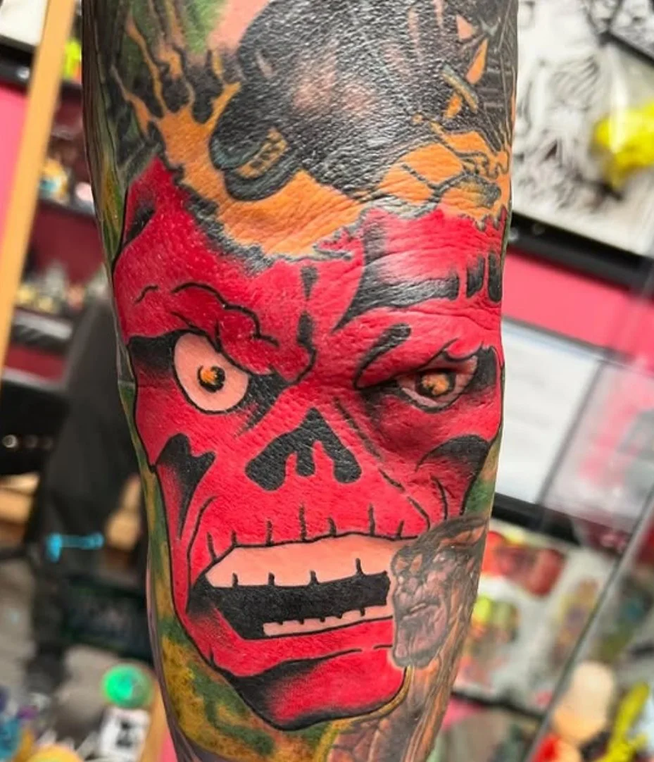 Colorful tattoo of a red skull with split features, one side a traditional skull and the other a zombie-like face, on a person's arm.