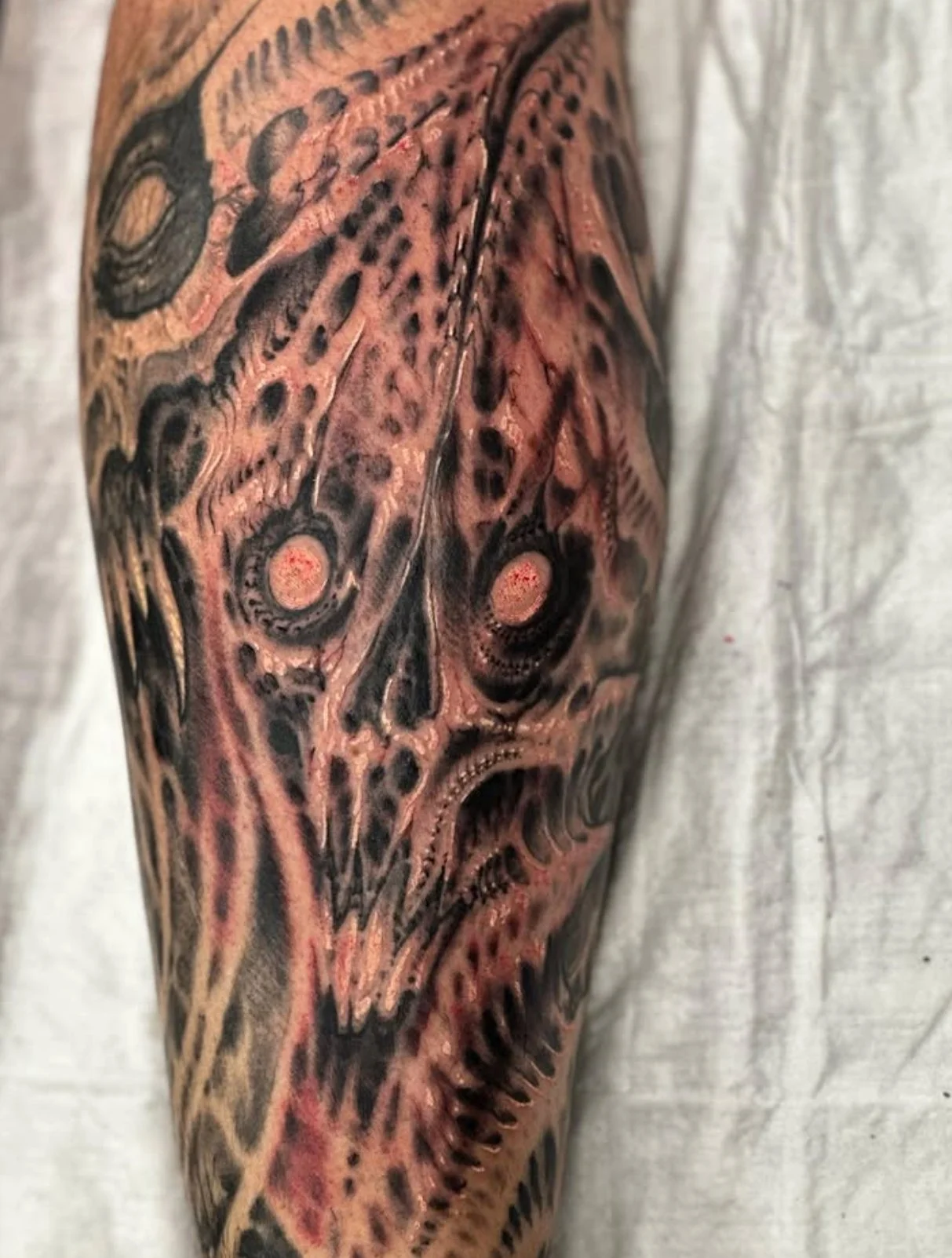 Close-up of a person's arm featuring a detailed black and gray tattoo of a fierce skull face with sharp teeth and textured scales.