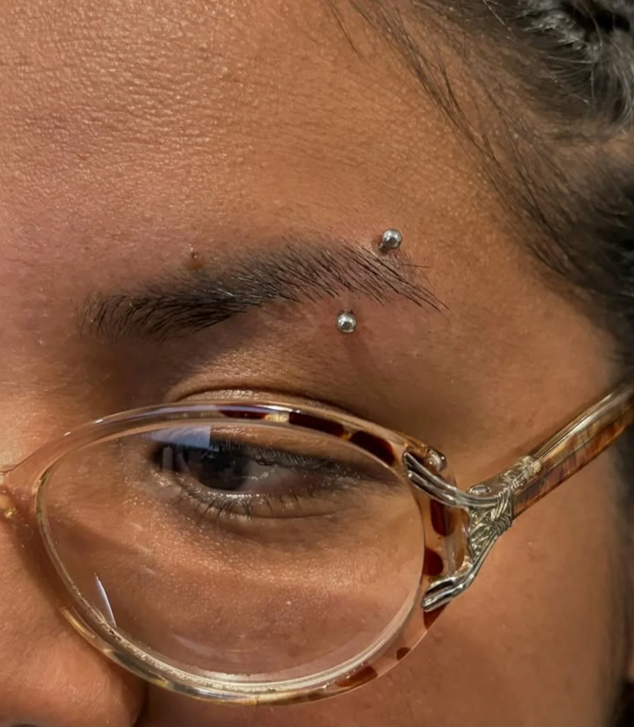 Close-up of a person’s face showing eyebrow piercings above the right eye and wearing tortoiseshell glasses.