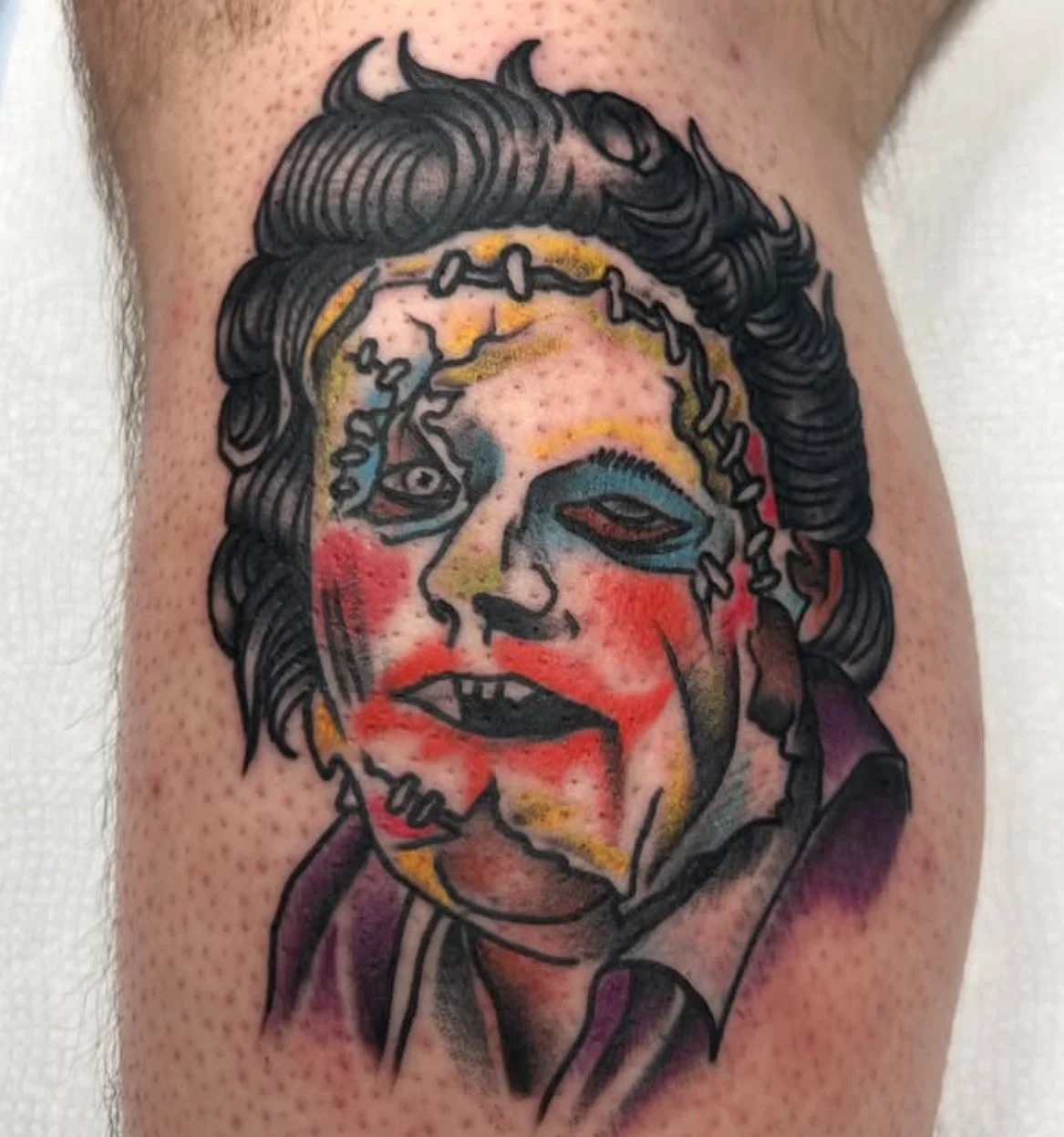 Colorful tattoo of a Leatherface mask with woman's face with expressive makeup, loose hair, and a chain around her head, in a traditional style.