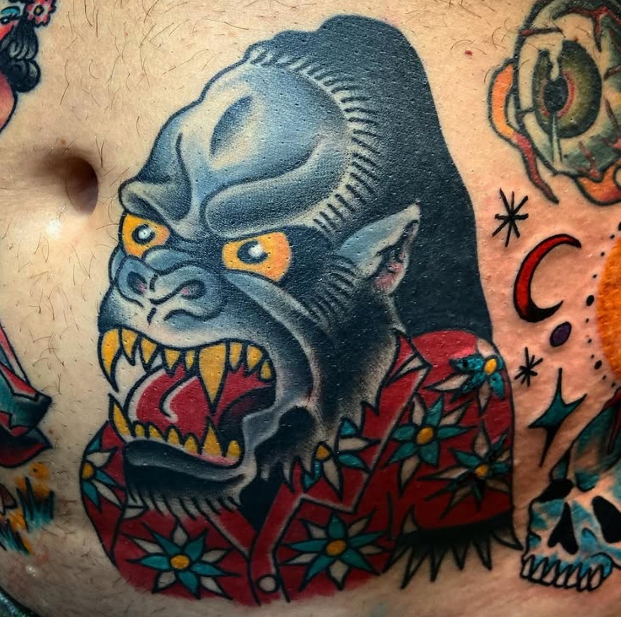 Colorful tattoo of a snarling ape with yellow eyes, sharp teeth, and a red floral shirt, surrounded by smaller celestial-themed symbols.