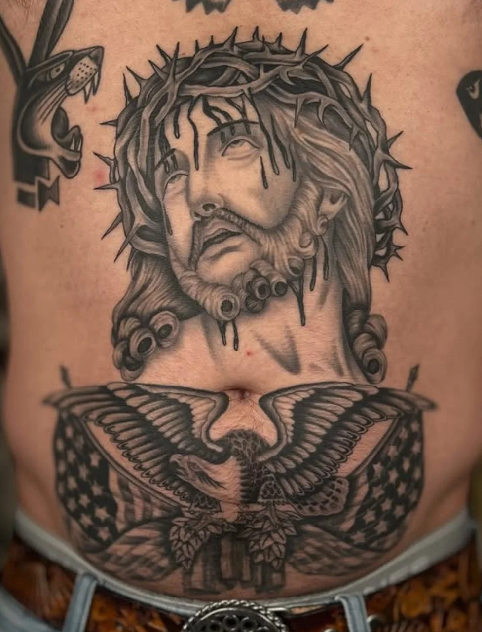Tattoo of Jesus Christ with a crown of thorns, long hair, and a beard, with a sad expression. Below, a large butterfly with American flag pattern on its wings.