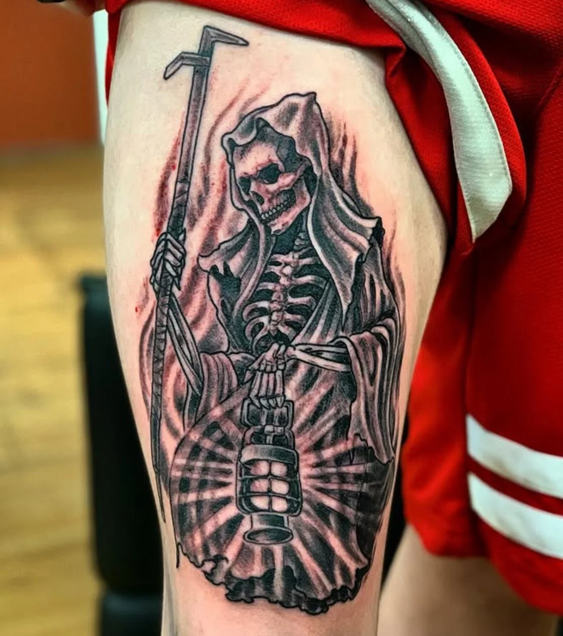 Tattoo of a skeleton with a hood, holding a scythe and a grenade, on a person's upper arm.