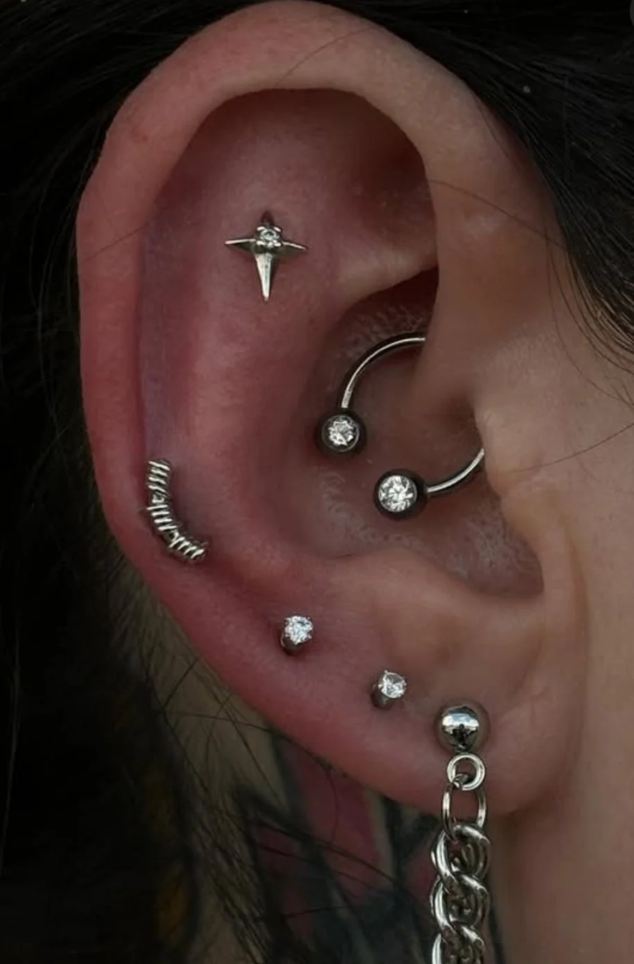 Close-up of a person's ear showing multiple piercings with various earrings, including a star-shaped stud, a hoop with black accents, small rhinestone studs, and a dangling chain-style earring.
