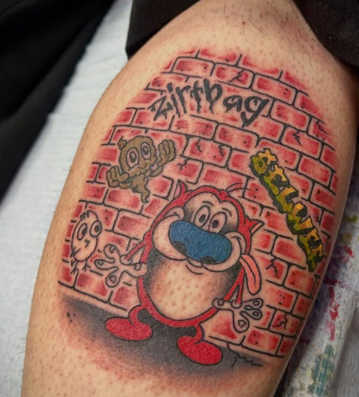 Colorful tattoo of Stimpy from Ren and Stimpy, surrounded by graffiti-style text and characters, on upper arm with brick wall background.
