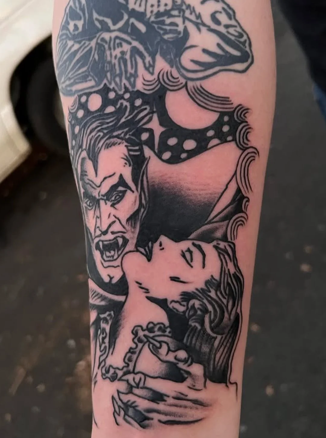 Tattoo of a fierce-looking Dracula with long hair and an angry expression, yelling at a woman with long hair and closed eyes, with one hand gripping her arm.