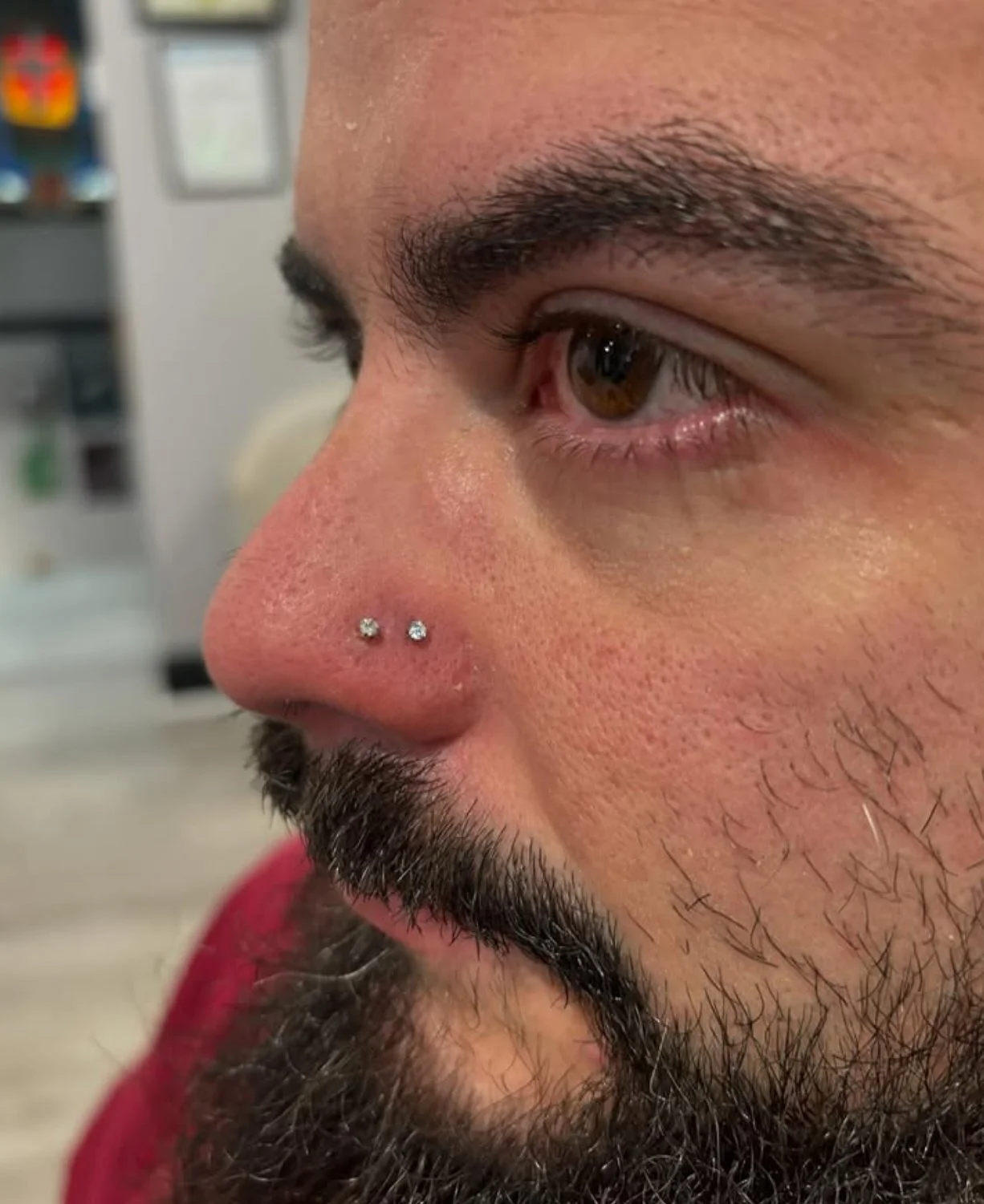 Close-up of a man's face focusing on his nose with two small nose piercings and his brown eye, along with his dark beard and mustache.
