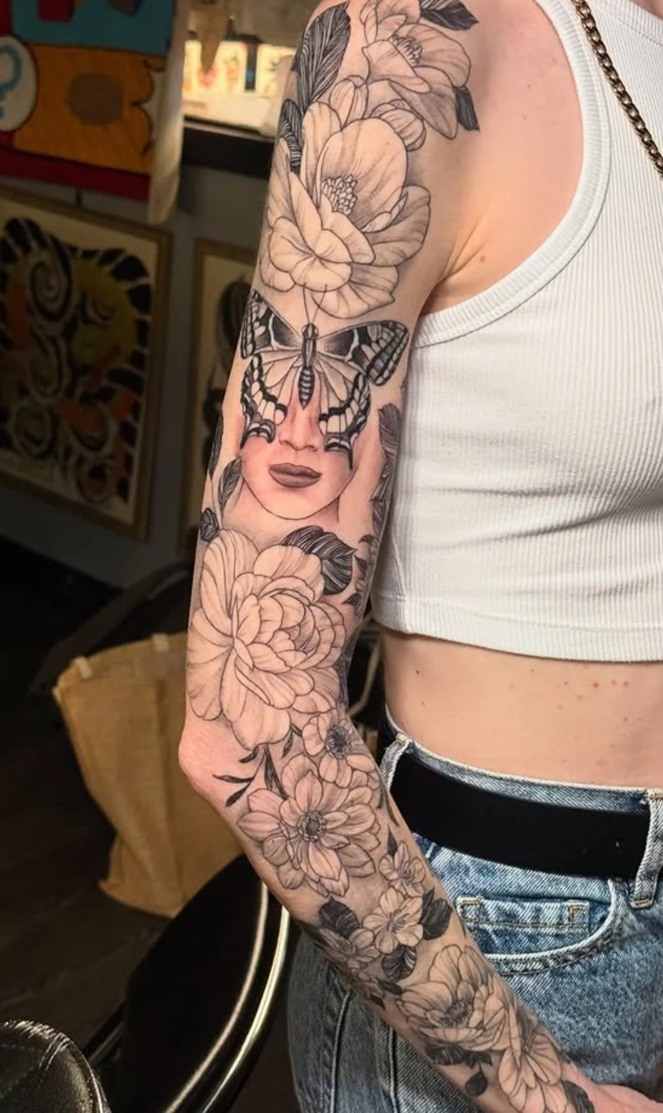 Tattoo sleeve on a person's arm featuring large flowers, a woman's face, and a butterfly, with the person wearing a white sleeveless crop top and blue jeans.