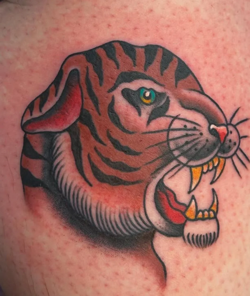 Tattoo of a tiger's face with detailed fur, sharp teeth, and a colorful eye.