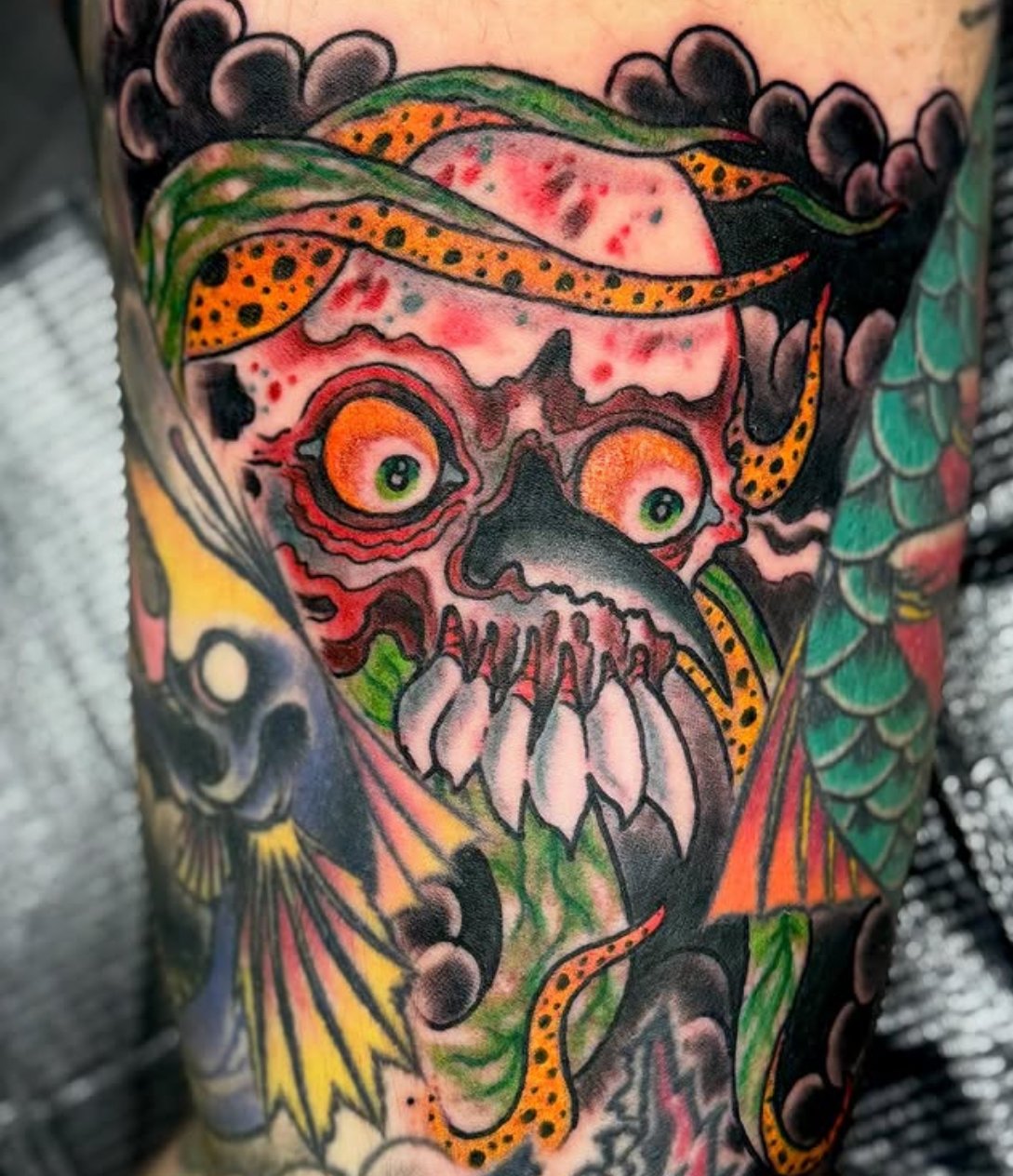 A colorful tattoo of a skull with exaggerated eye sockets, surrounded by snakes, scales, and other vivid imagery.