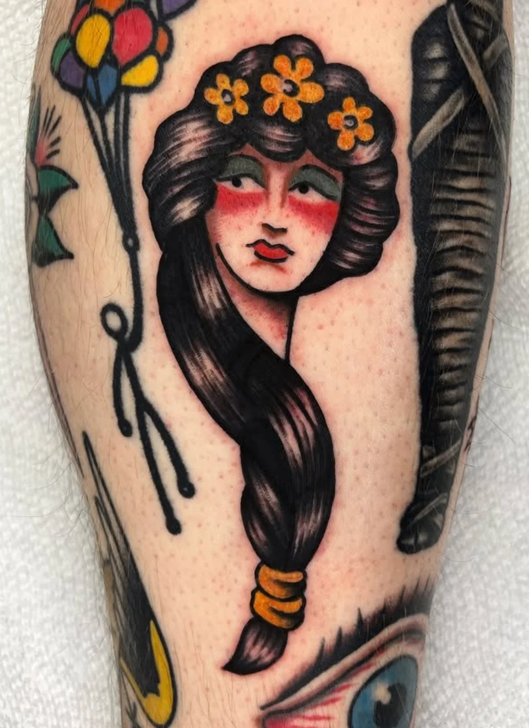 Tattoo of a woman with dark hair, red cheeks, and a crown of orange flowers, in traditional style.