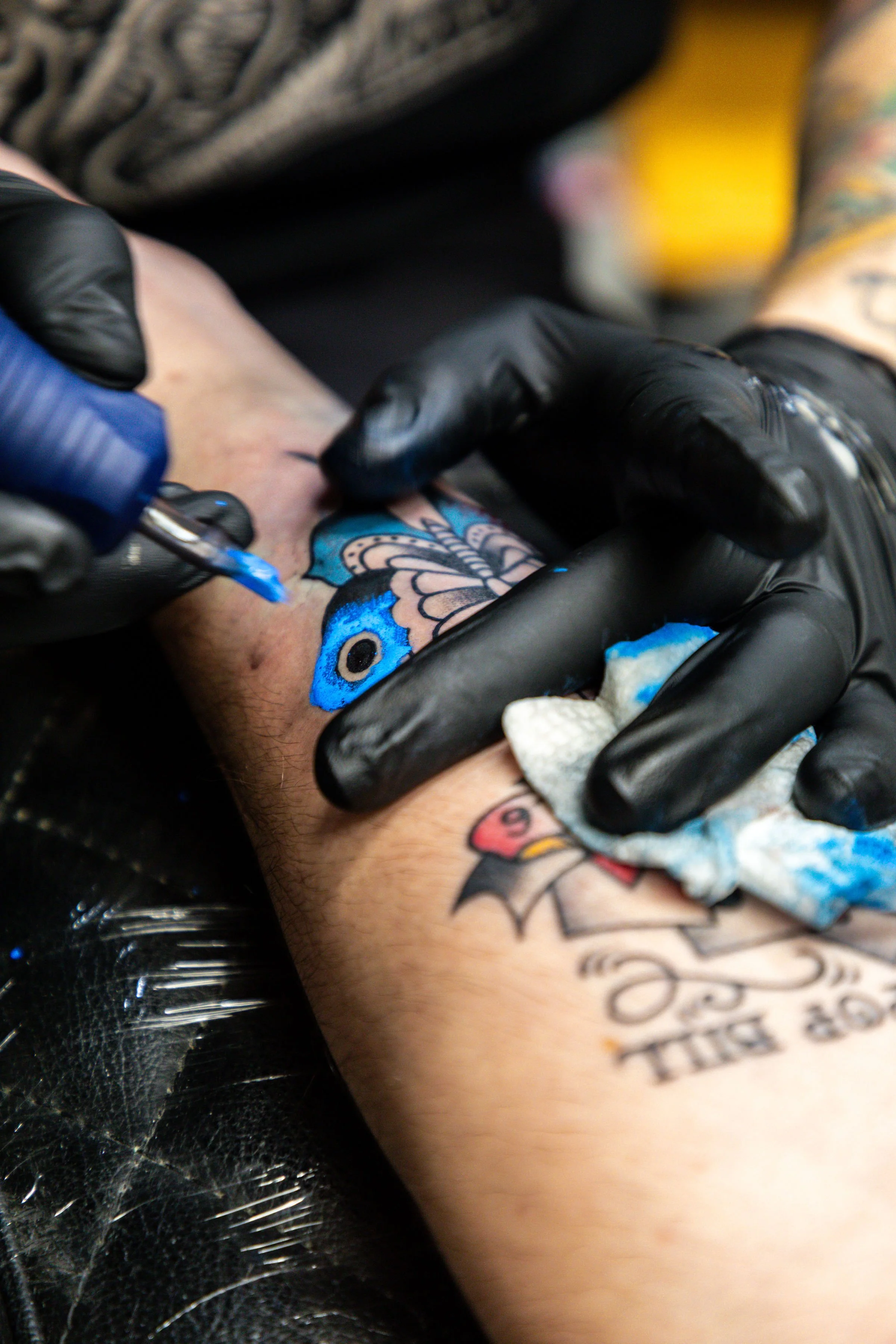 A person getting a colorful butterfly tattoo on their arm at a tattoo studio. The tattoo artist is using a tattoo gun, wearing black gloves, and using a cloth to wipe excess ink.