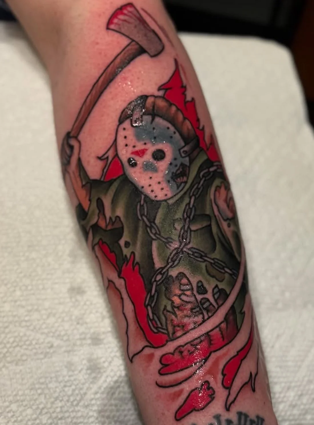Tattoo of Jason Voorhees from Friday the 13th, depicted as a hockey mask with a red slash and wearing a chain, on an arm with red and black flesh rips in the background.