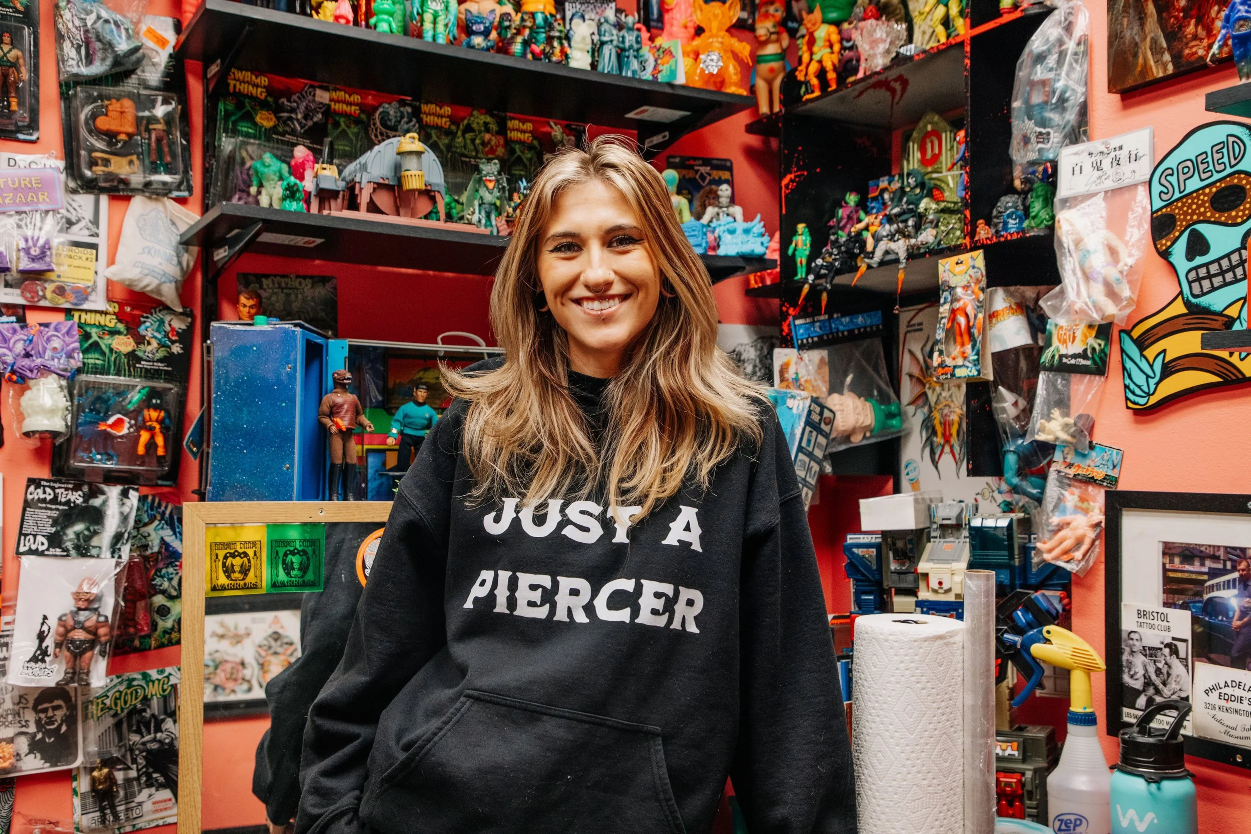 A young woman with wavy dirty blonde hair smiling in a toy or collectibles store, surrounded by action figures, toys, and pop culture memorabilia on shelves and walls.