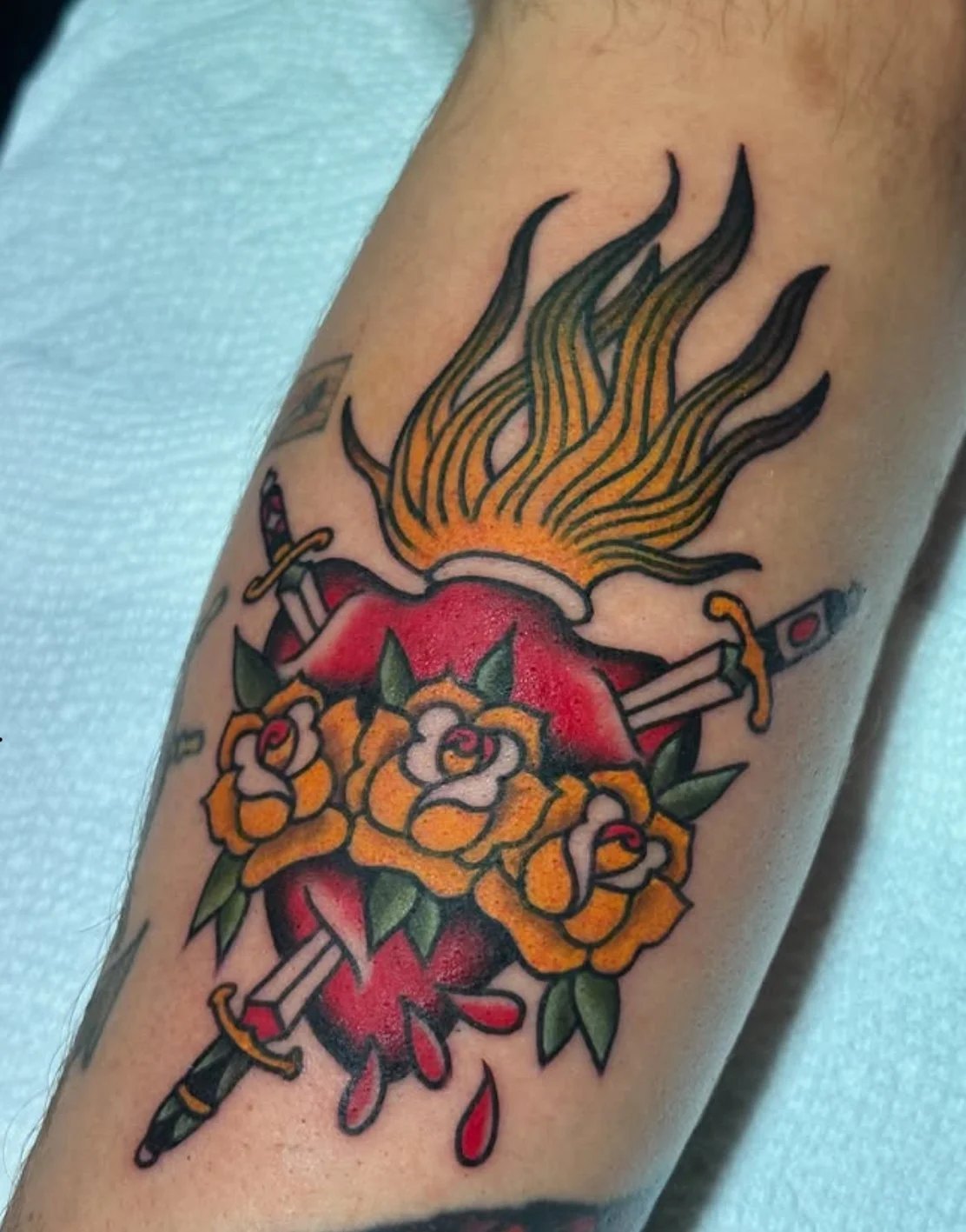 Tattoo of a sacred heart with flames, roses, and swords in traditional tattoo style.