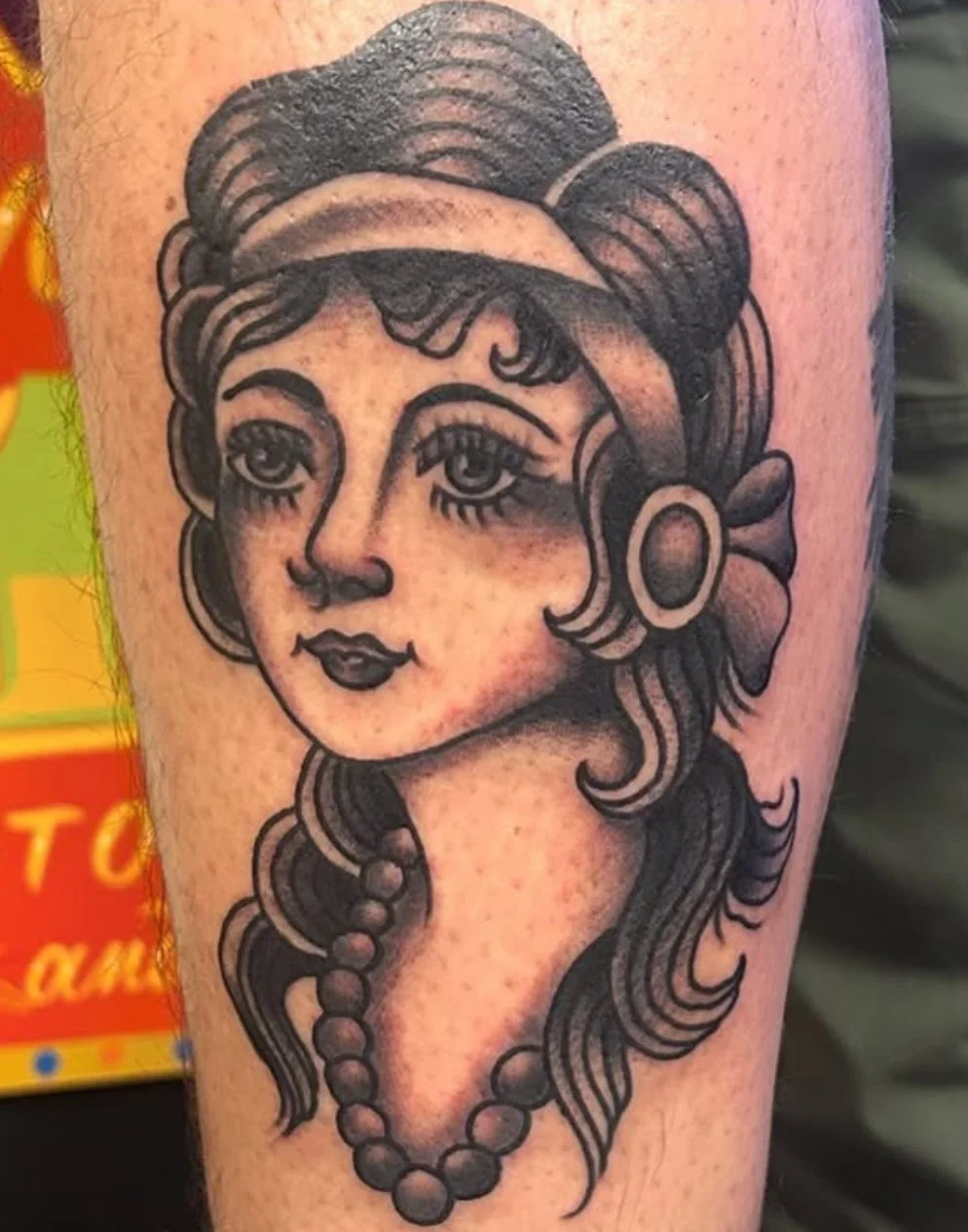 Tattoo of a vintage-style woman with detailed eyes, curly hair adorned with a bow and large circular earrings, wearing a beaded necklace.