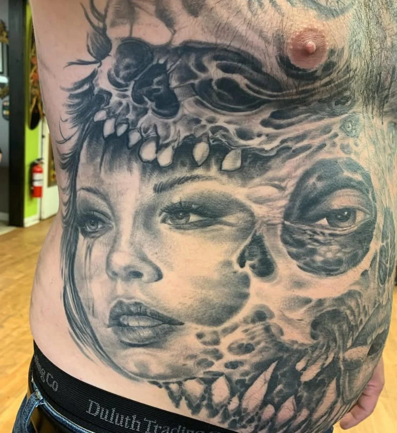 Black and gray tattoo on a person's torso depicting a woman's face with detailed eyes and lips, blending into abstract and animal-like features.