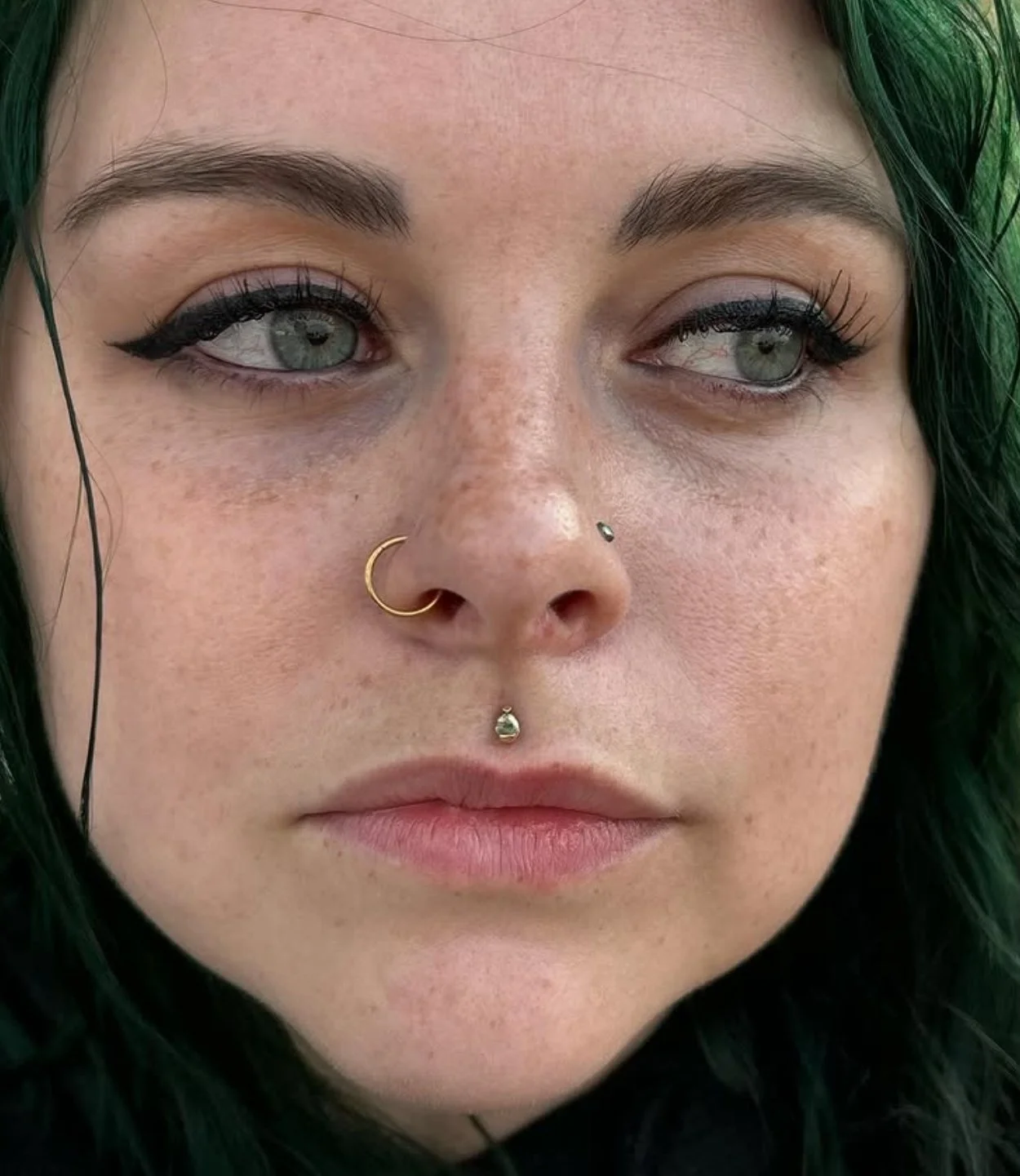 Close-up of a woman's face with green hair, wearing multiple facial piercings, including a septum piercing with a gold ring, a nostril piercing, and a labret with a small gem, and with makeup featuring winged eyeliner and natural lipstick.