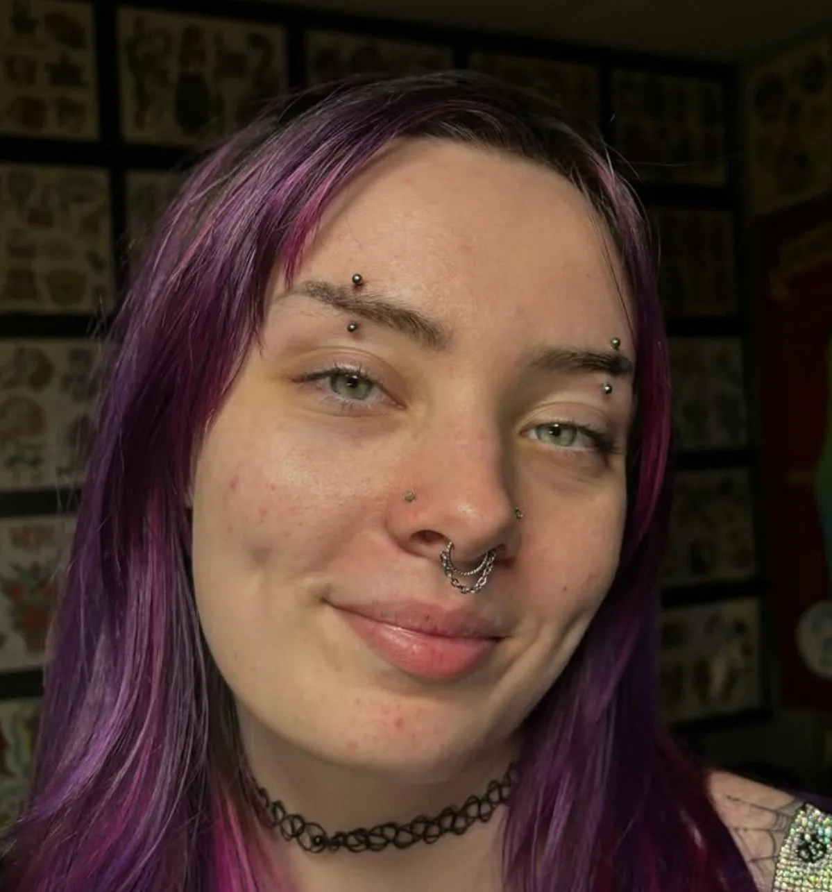 A young woman with purple hair and multiple facial piercings, including nose, eyebrow, and lip piercings, smiling in front of a tattoo flash wall.