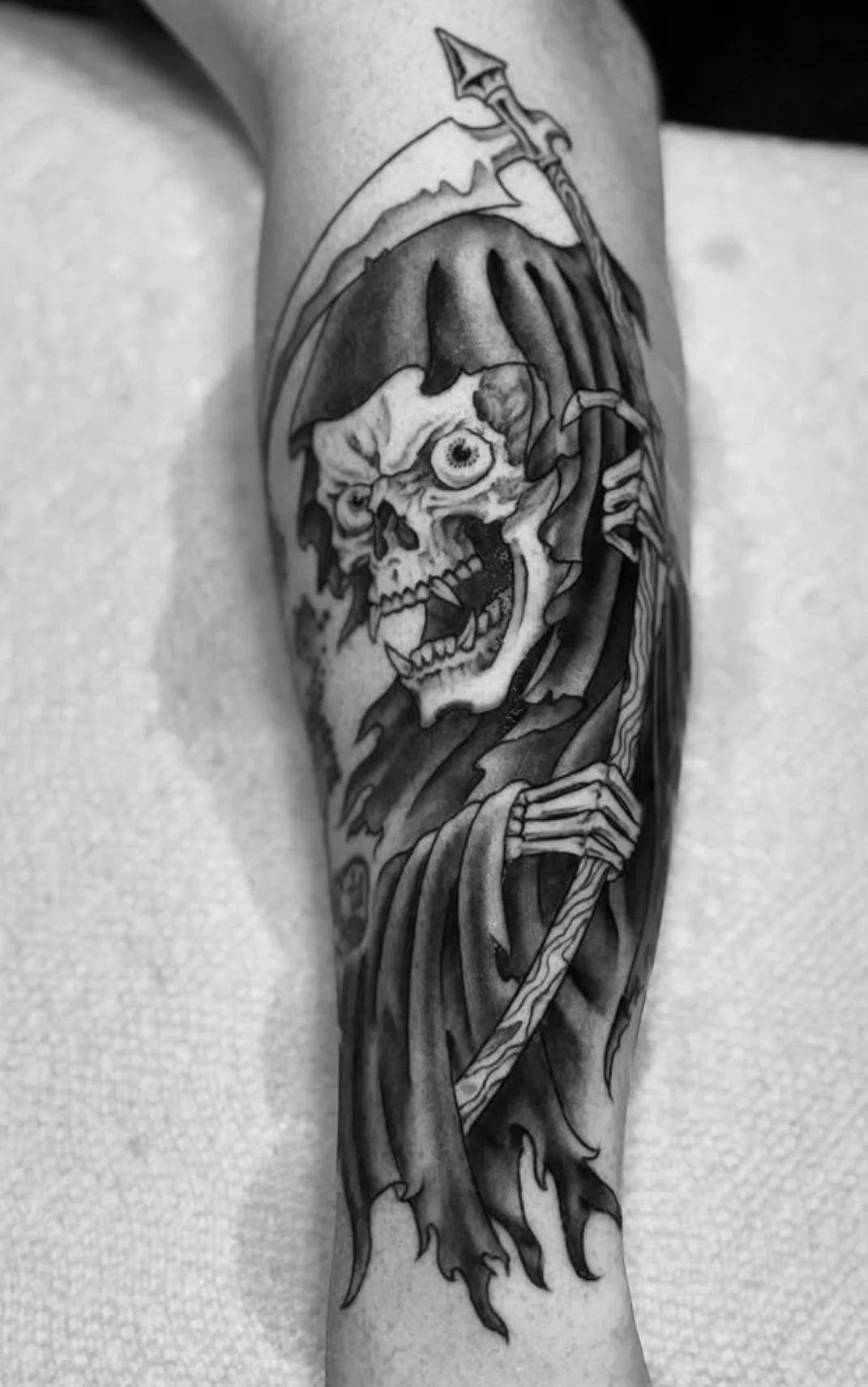 Black and gray tattoo of a skeletal Grim Reaper, holding a scythe, on a persons forearm.