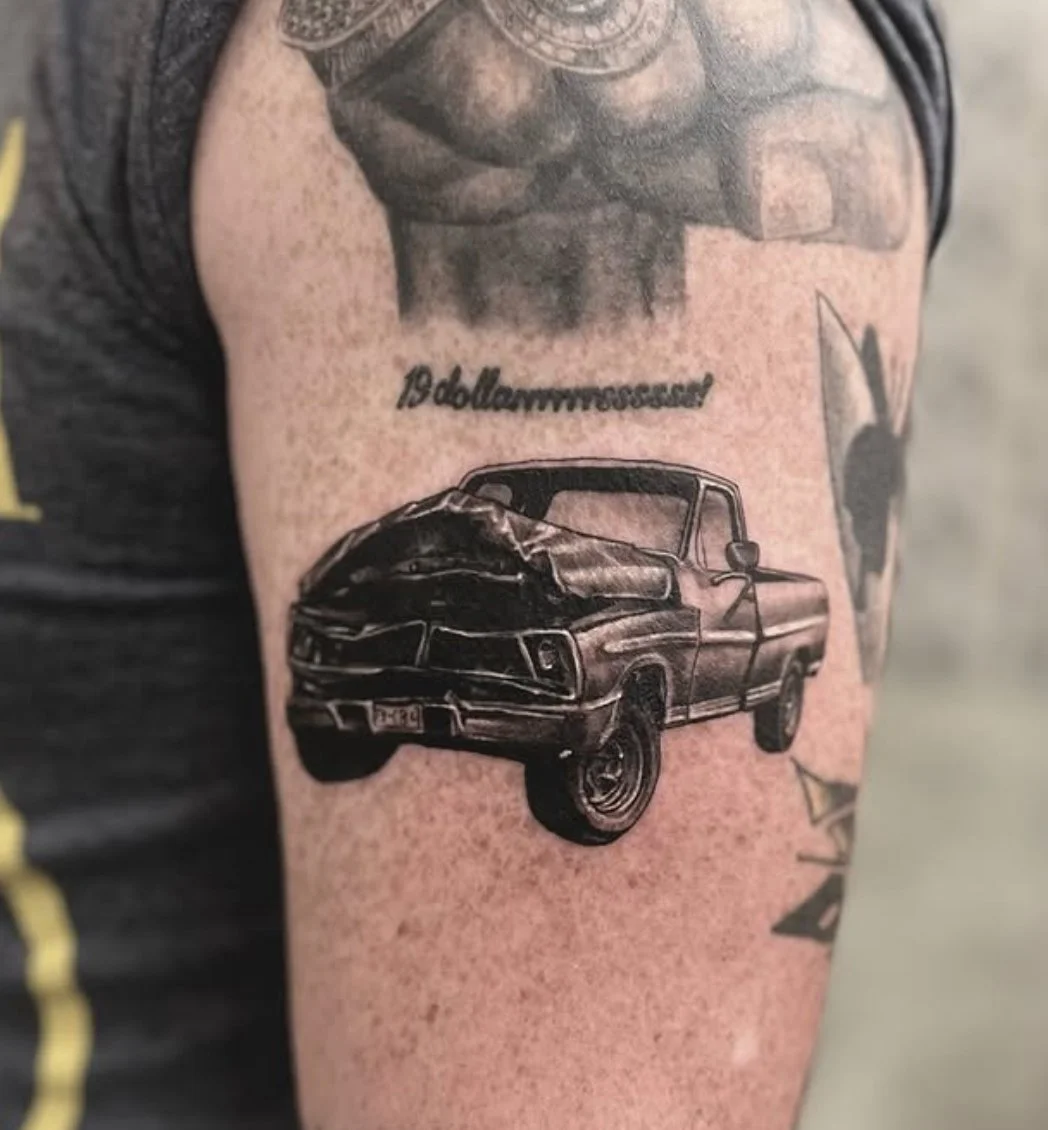 Tattoo of a damaged pickup truck from Post Malone’s album with text above it that reads '19 dollarrrrrrssssss' on a person's upper arm.