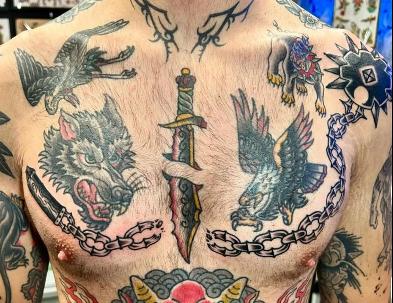 Tattooed chest featuring a central dagger with decorative elements, a snarling black wolf and an eagle, chains, and other traditional-style tattoo imagery.