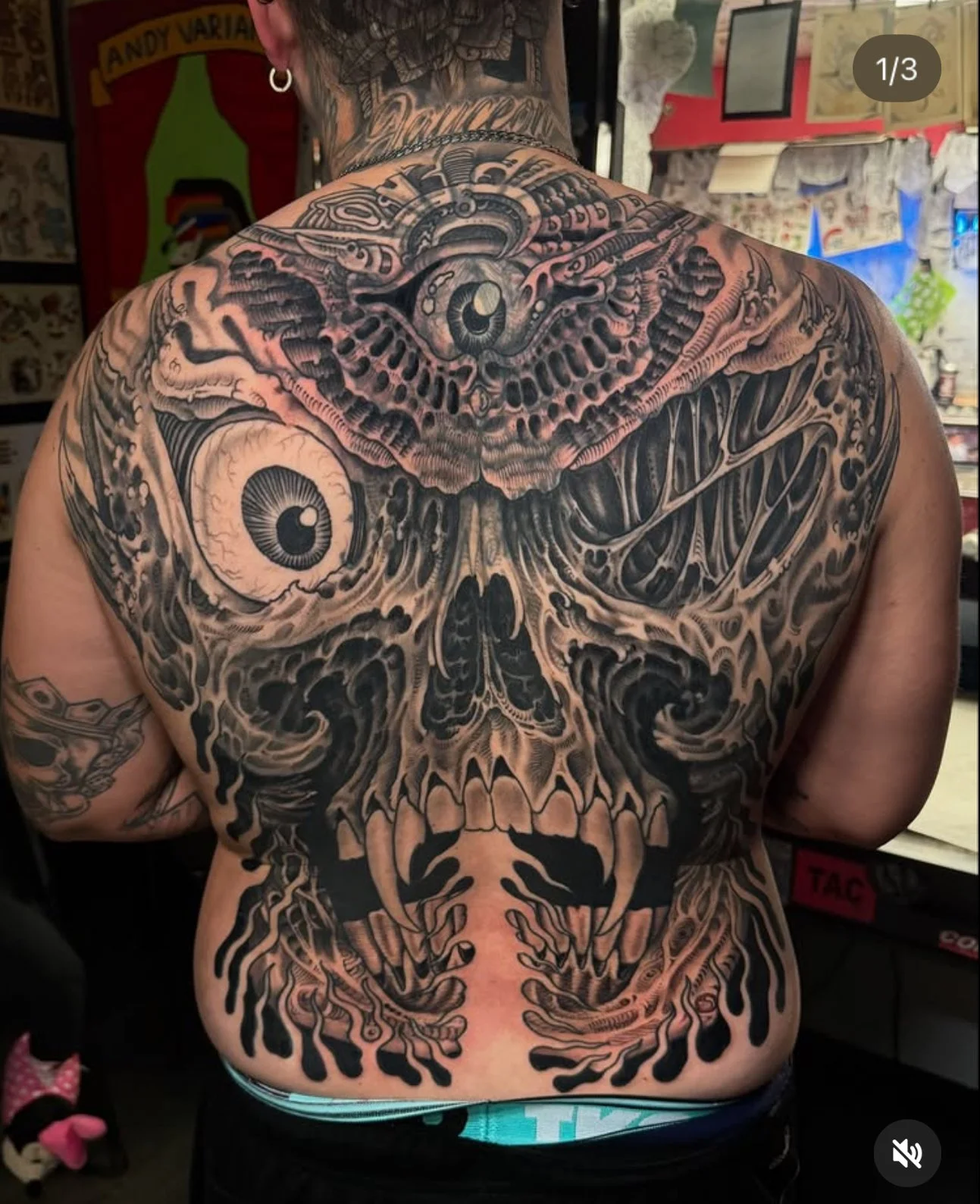 The person's back features a large, detailed tattoo of a skull with eyes, surrounded by intricate designs, dark shading, and bold lines.