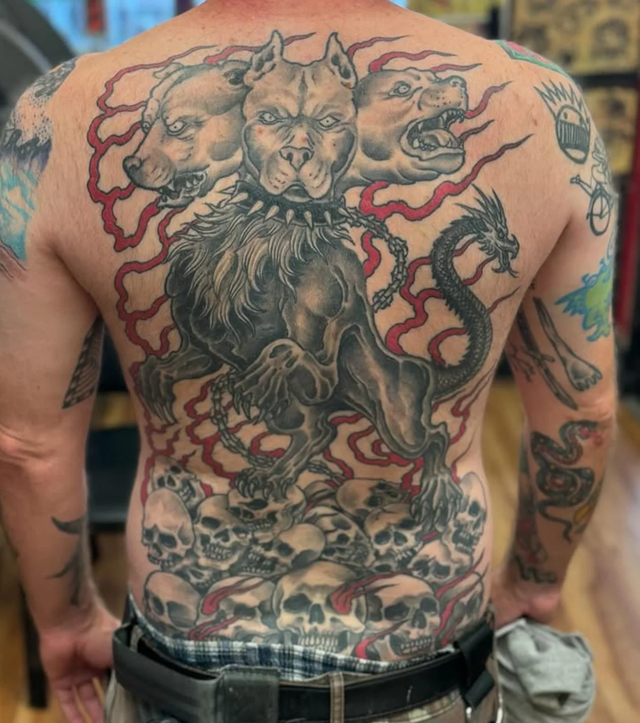 Tattoo of a three-headed dog, a Cerberus, a lion, and a snake on a person's chest, with skulls and red swirling lines.