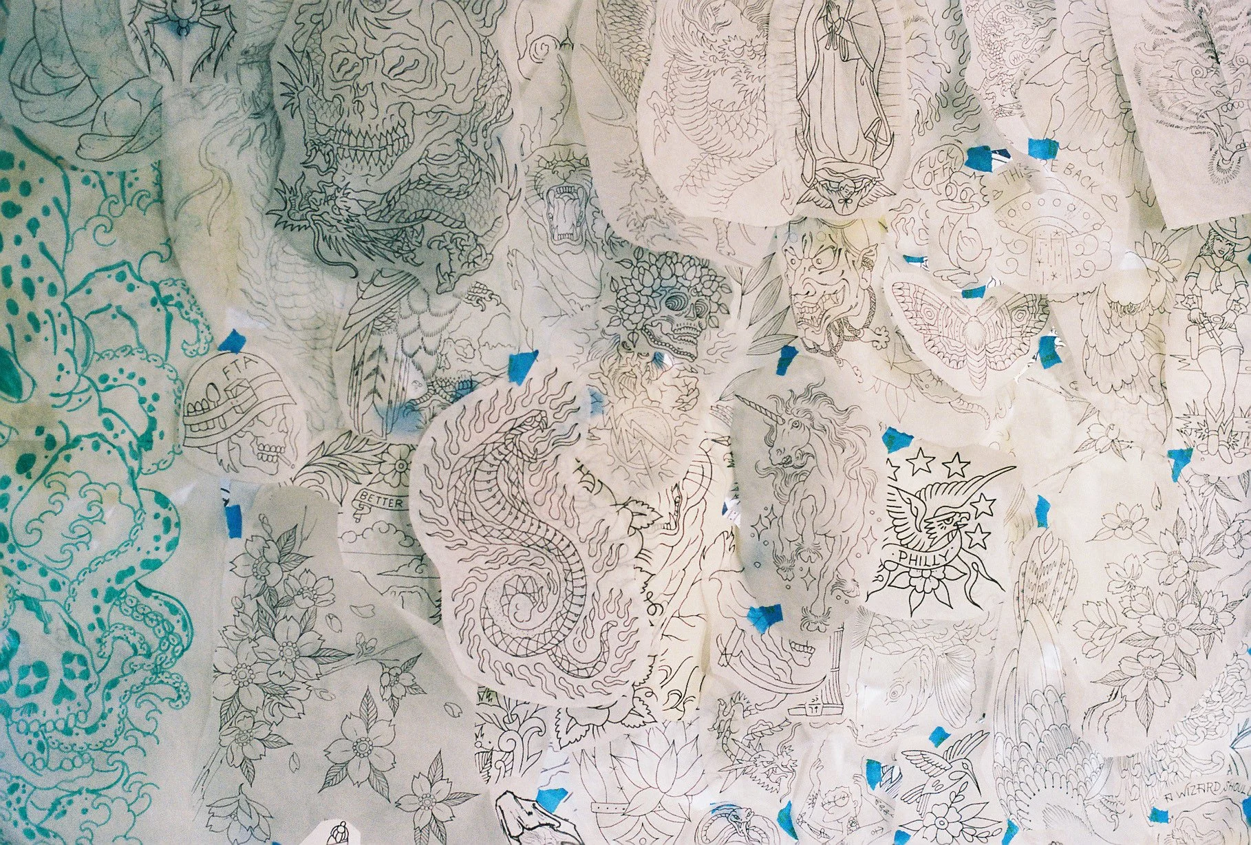 Collection of black and white tattoo stencils taped on a wall, featuring various designs including animals, mythical creatures, and floral patterns.