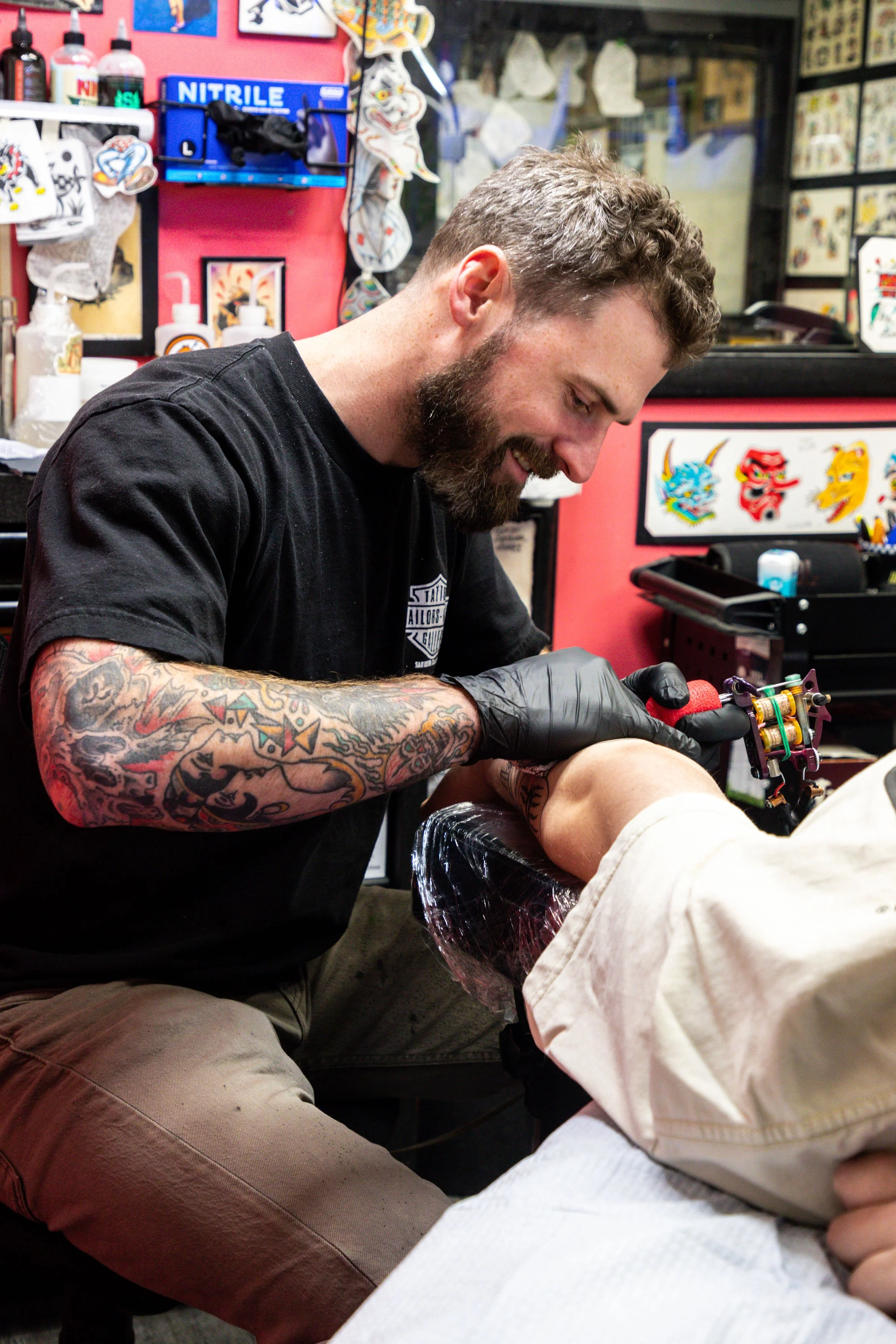 A man with a beard and tattooed arms is tattooing a person's arm in a tattoo shop. The background has tattoo-themed artwork and supplies.