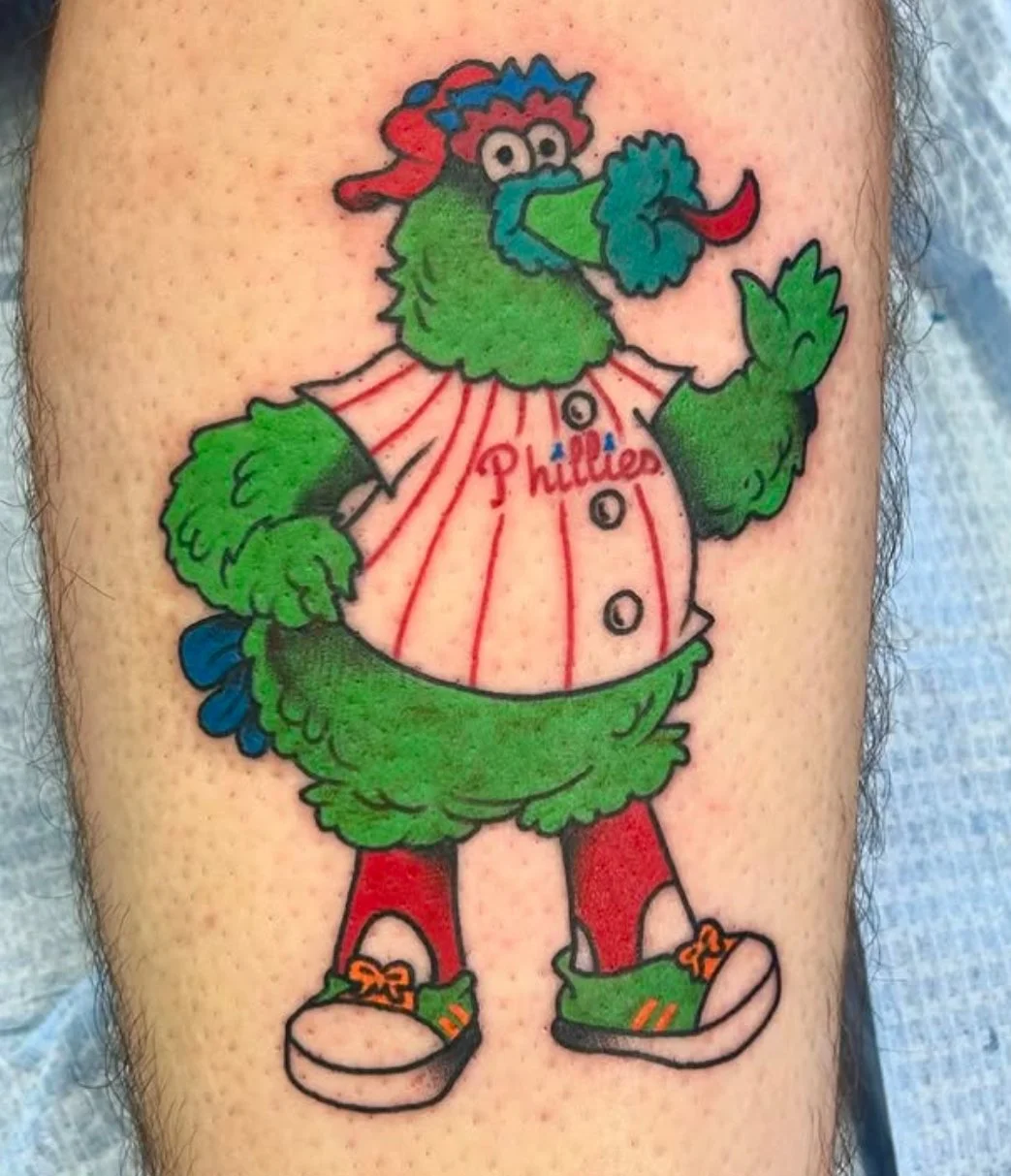 Tattoo of the Philly Phanatic in a Phillies baseball jersey, wearing red socks and green sneakers, with a cheerful pose.