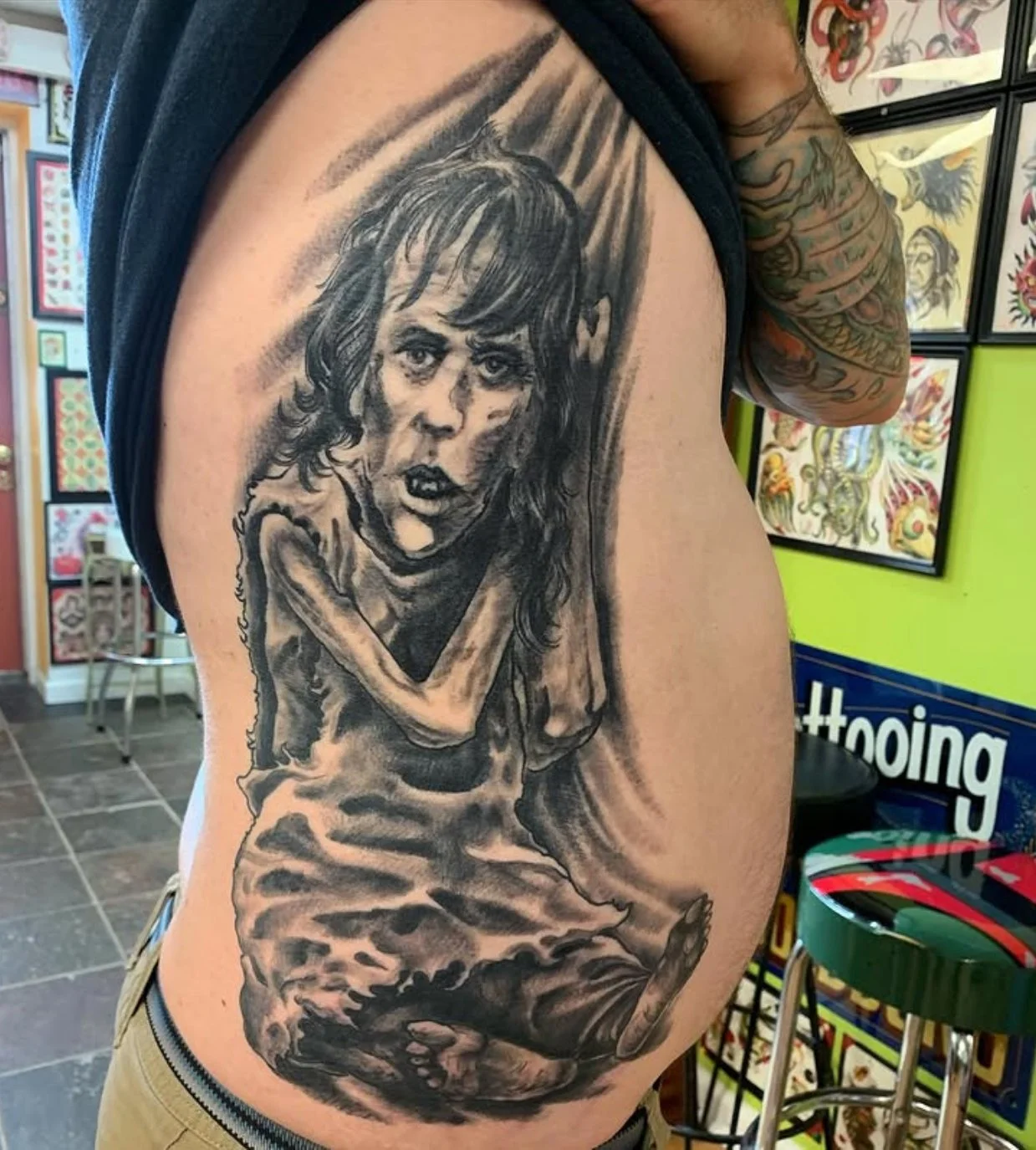 Black and gray tattoo of a young girl with curly hair and an expressive face, sitting cross-legged with arms resting on her knees, covering her mouth in shock or horror.