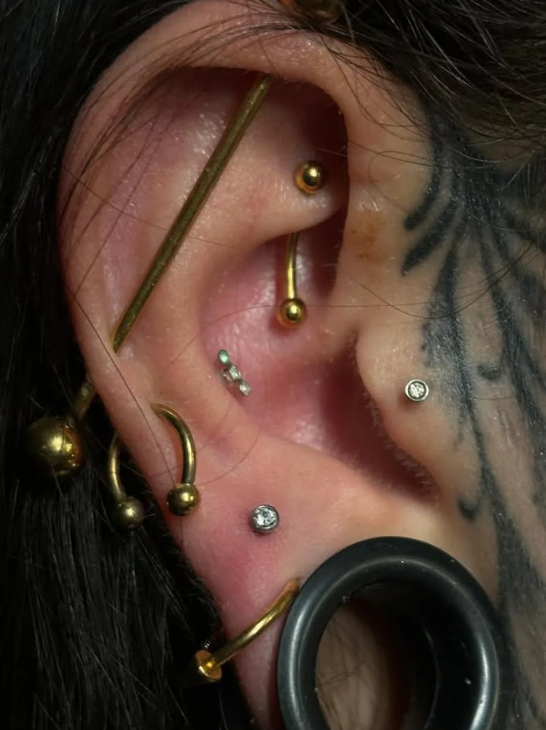 Close-up of a person's ear with multiple piercings, including helix, tragus, and lobe, adorned with gold and diamond earrings, and head tattoo in black ink.