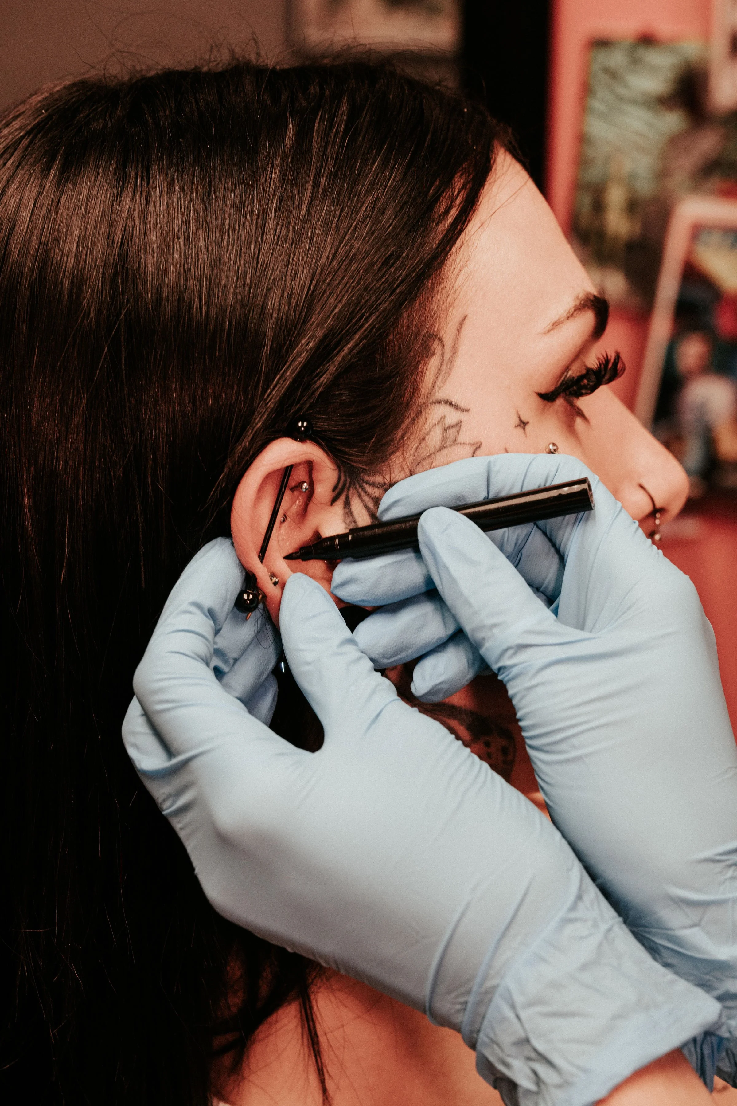 A person with tattoos on their face receives a piercing in their ear from a tattoo artist wearing blue gloves.