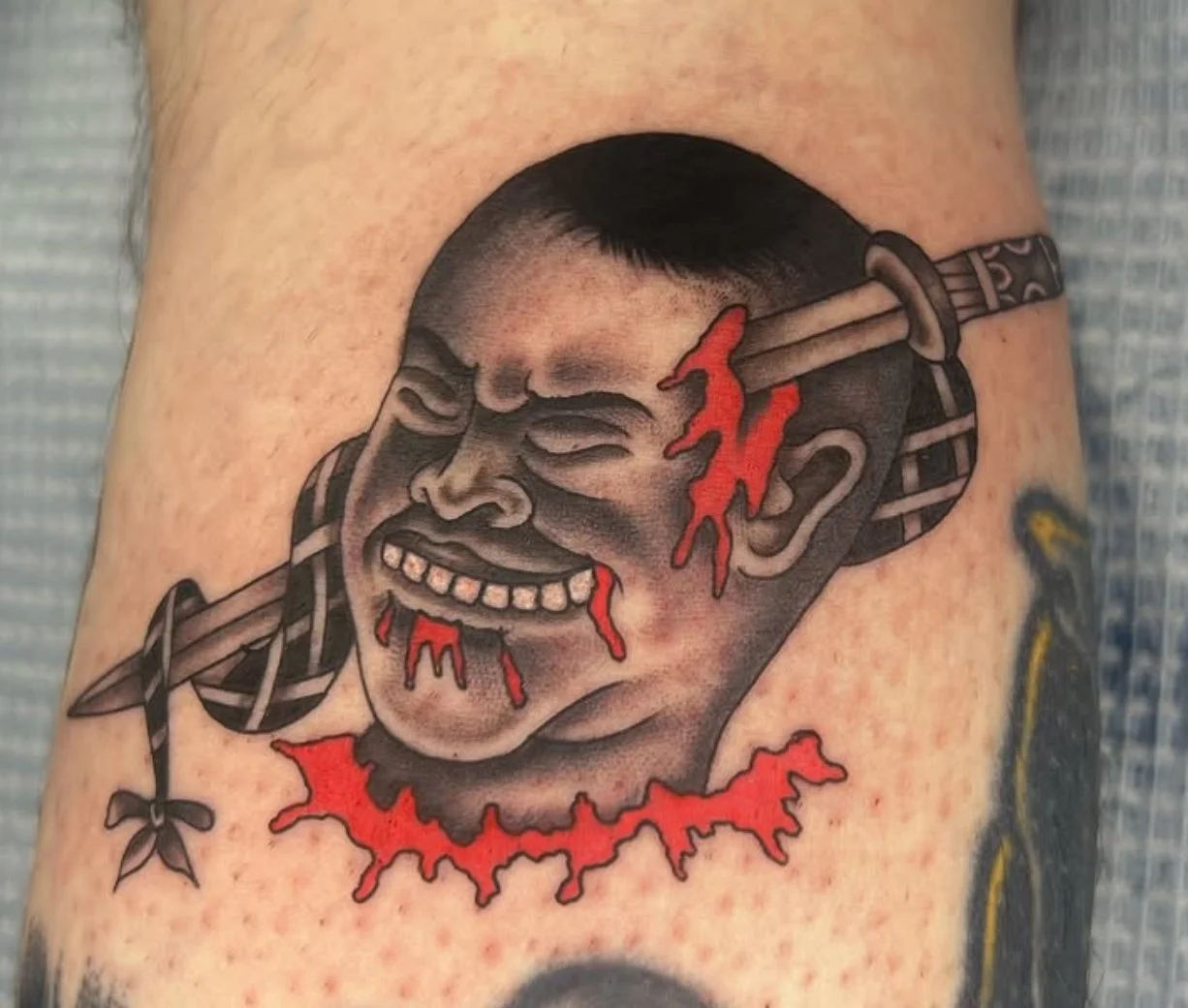 Tattoo of a crying, smiling man's face with a knife through his head and blood dripping, with a red bloody collar and blood dripping from his chin.