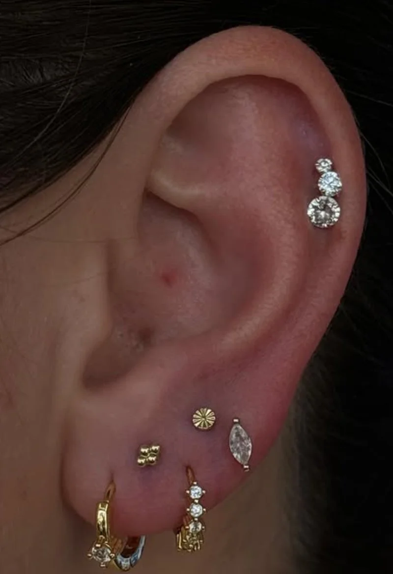 Close-up of a person's ear with multiple earrings, including a row of three sparkling diamond-like earrings and a gold hoop earring.