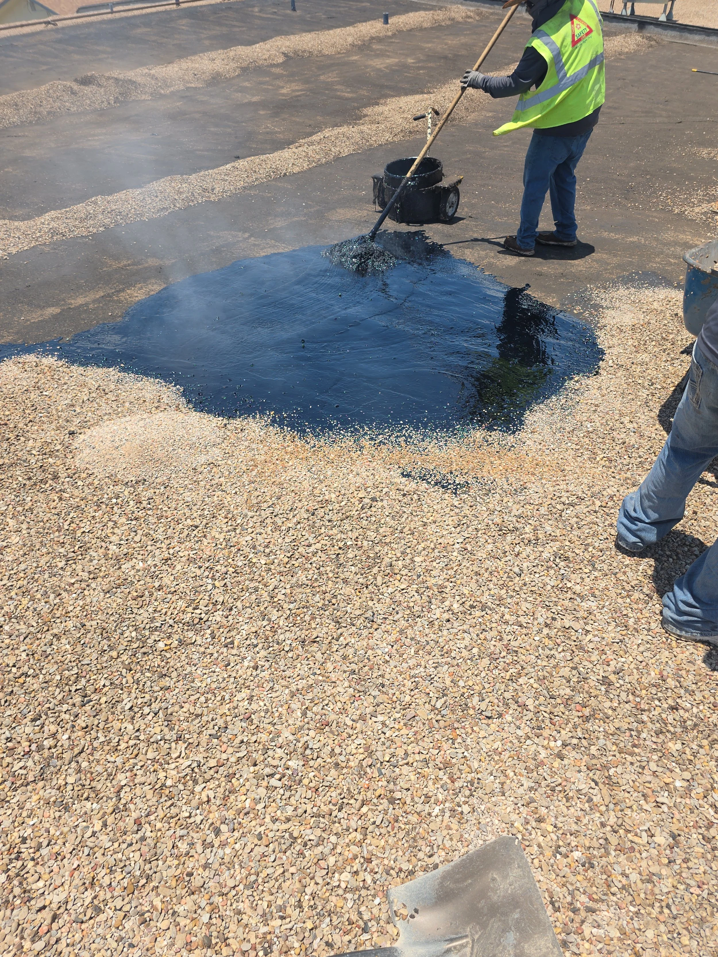 Worker in safety vest spreading black asphalt on a gravel surface using a rake, at a construction site.