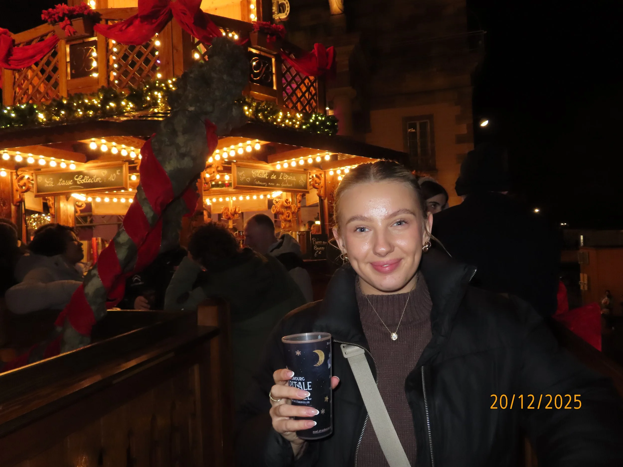 A young woman with blonde hair and earrings smiling at the camera, holding a black cup with a winter-themed design, at a festive outdoor event with Christmas decorations and lights, and a wooden stall in the background.