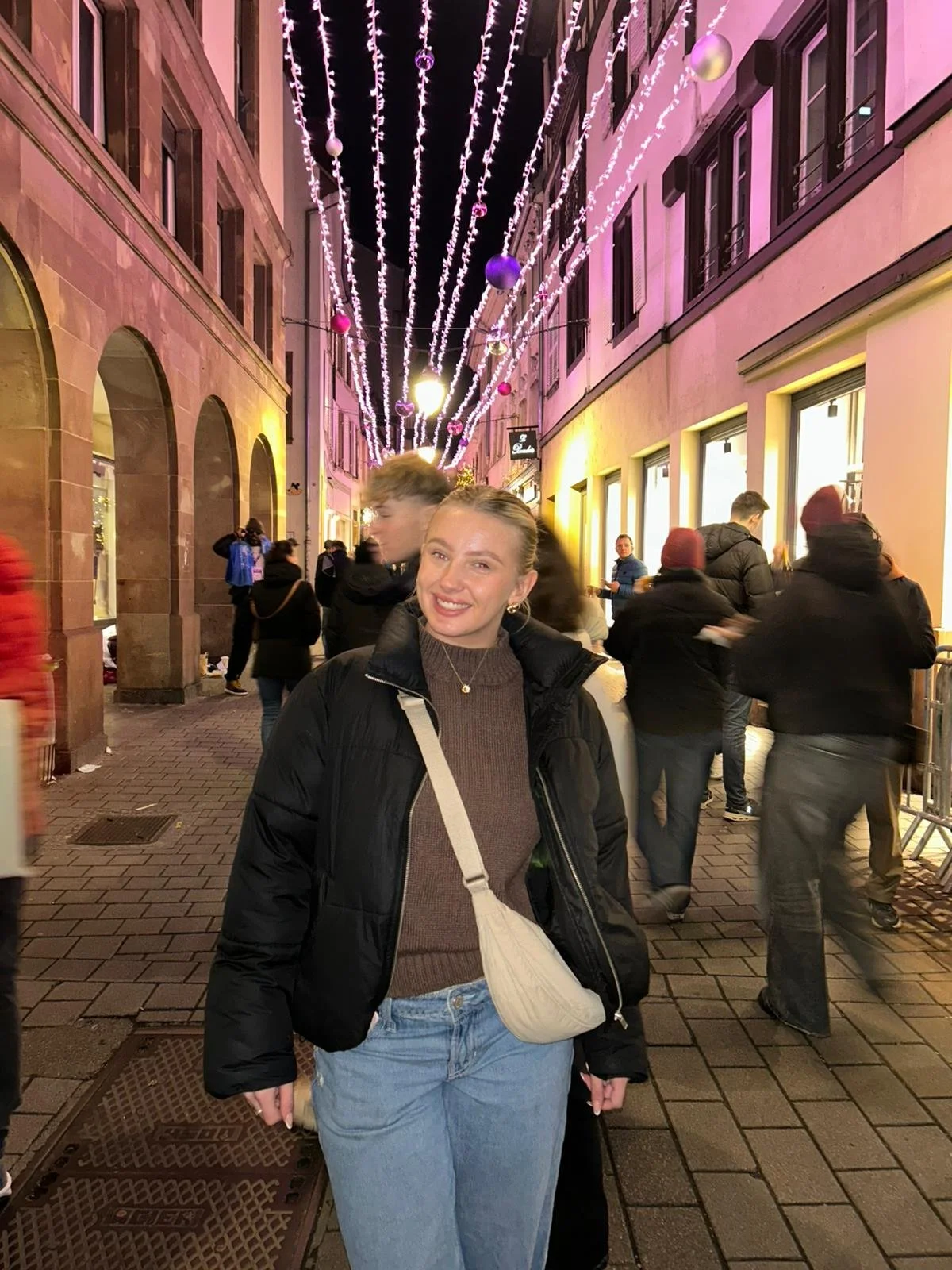 A young woman smiling at a nighttime street decorated with hanging string lights and ornaments, with people walking and shopping in the background.