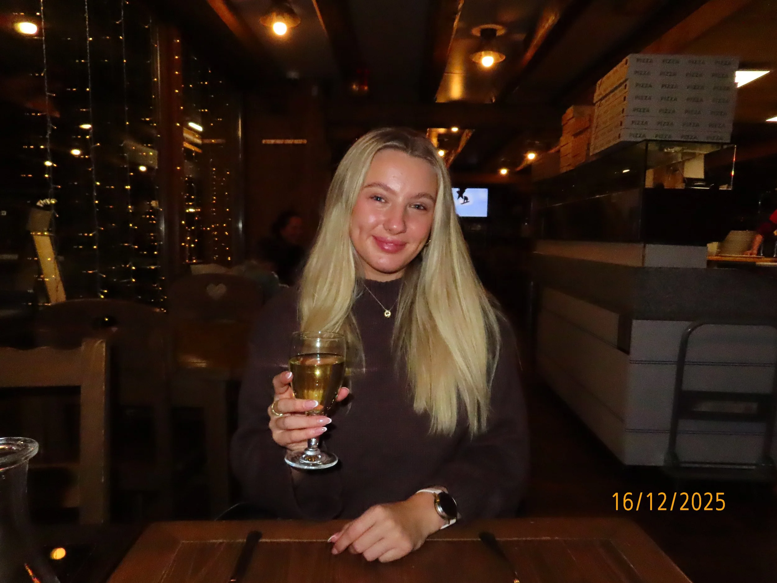 A young woman with long blonde hair, smiling, holding a glass of white wine, sitting in a dimly lit restaurant with a dark wooden ceiling and stacked pizza boxes in the background.