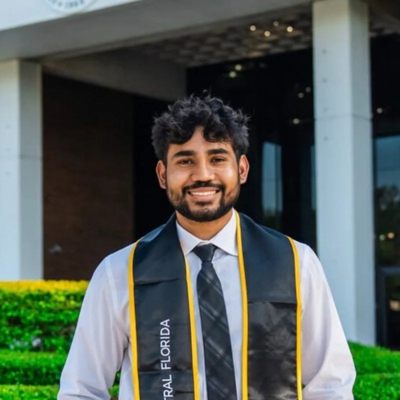A young man wearing a white shirt, a gray tie, and a graduation stole with 'Florida' written on it, standing outdoors in front of a modern building with greenery.