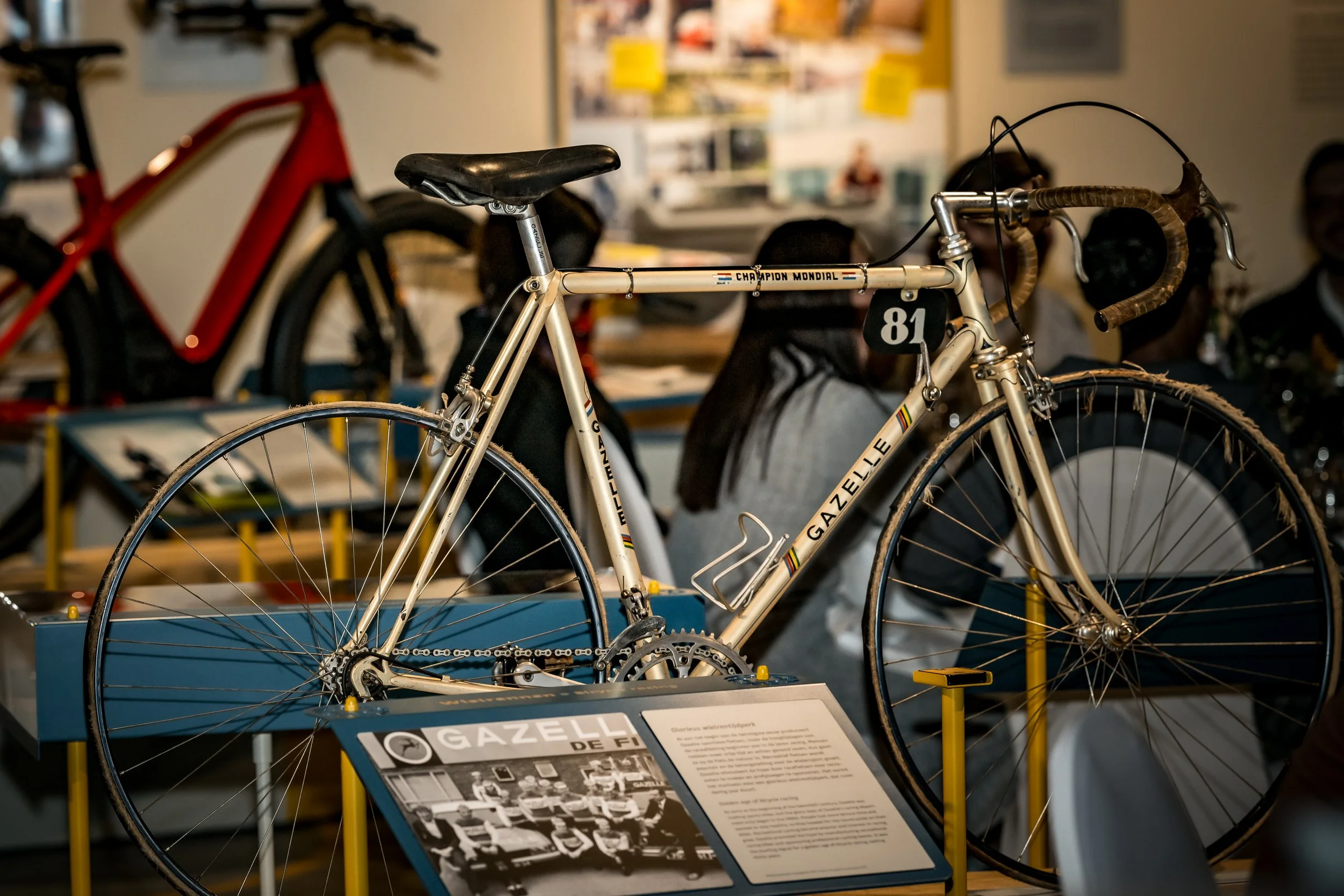 Een vintage gazell fiets op een display met informatief bord ernaast, in een museale omgeving met mensen op de achtergrond.