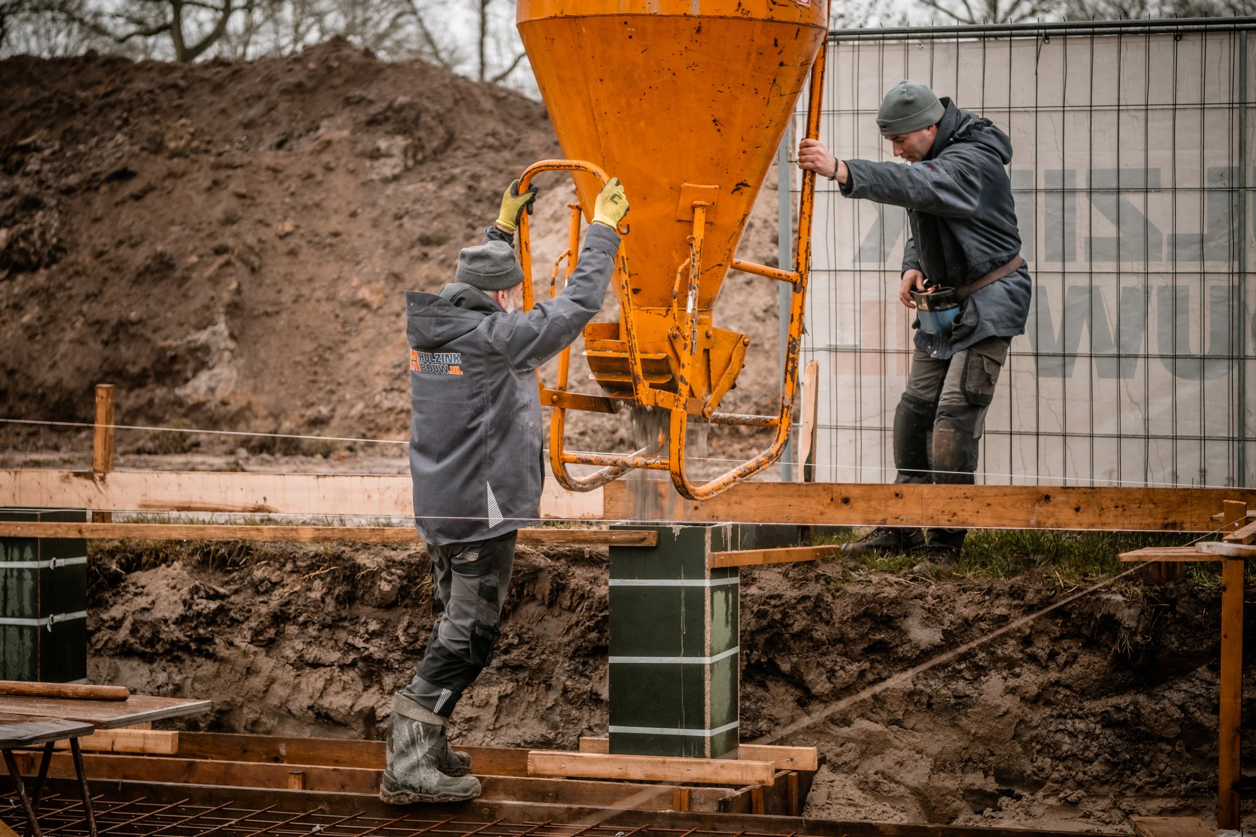 Twee bouwvakkers werken op een bouwplaats, een bedieningsmonteur bedient een mabine die betonnen funderingen schenkt terwijl een andere vakman toesnijdt en meet.
