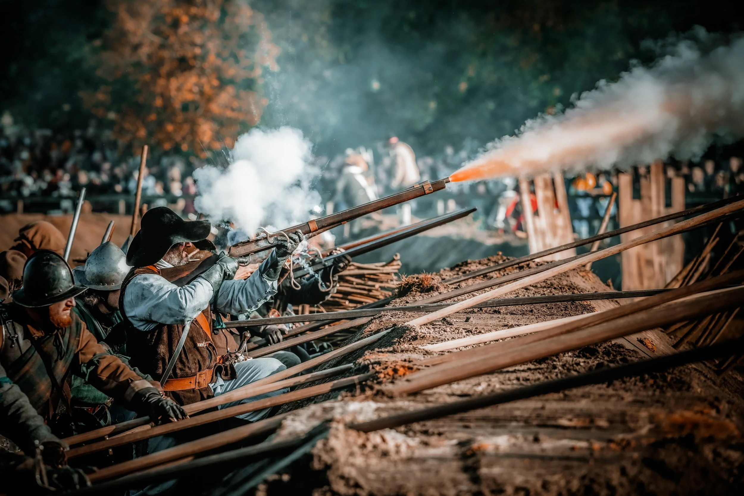 Historische veldslag met soldaten die musketen afvuren, in een veld met houten schotten en een bosrijke achtergrond.