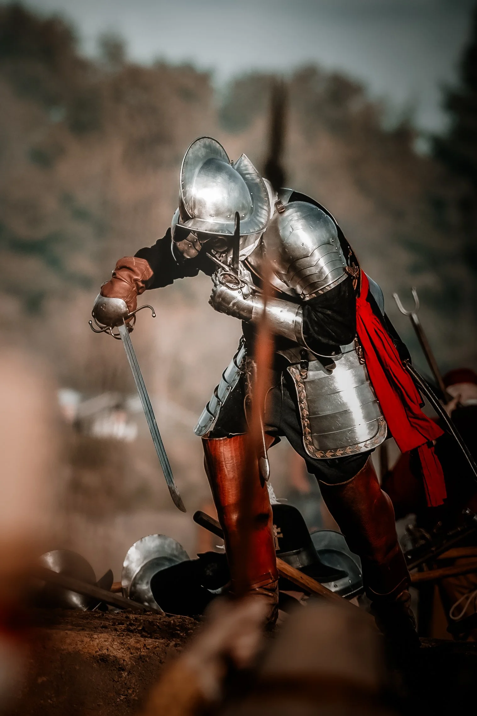 Een persoon gekleed in middeleeuwse metalen harnas en kleding, gehuld in heldere armor met een rode cape, tijdens een middeleeuws kledingspel of reenactment, buigt met een zwaard op een veld met een natuurrijke achtergrond.