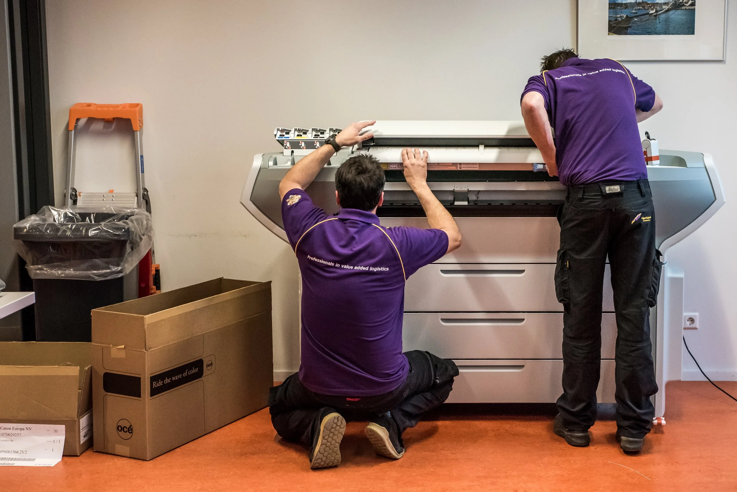 Twee technici in paarse uniformen repareren een groot multifunctioneel apparaat, mogelijk een printer of kopieermachine, in een kantoorruimte.