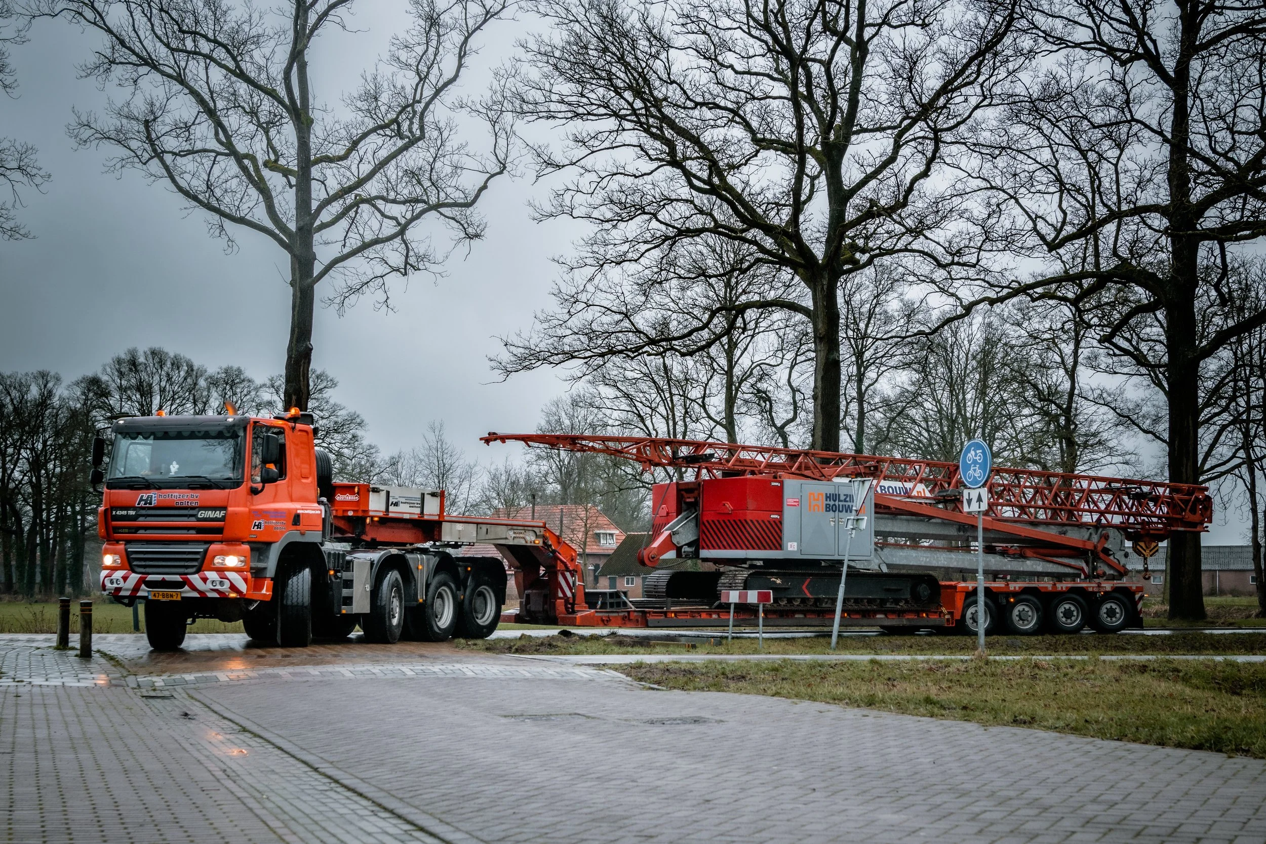 Grote rode vrachtwagen met uitgestrekte telescooparm en containeer op een parkeerplaats met bomen en een overwegpaal op de achtergrond.