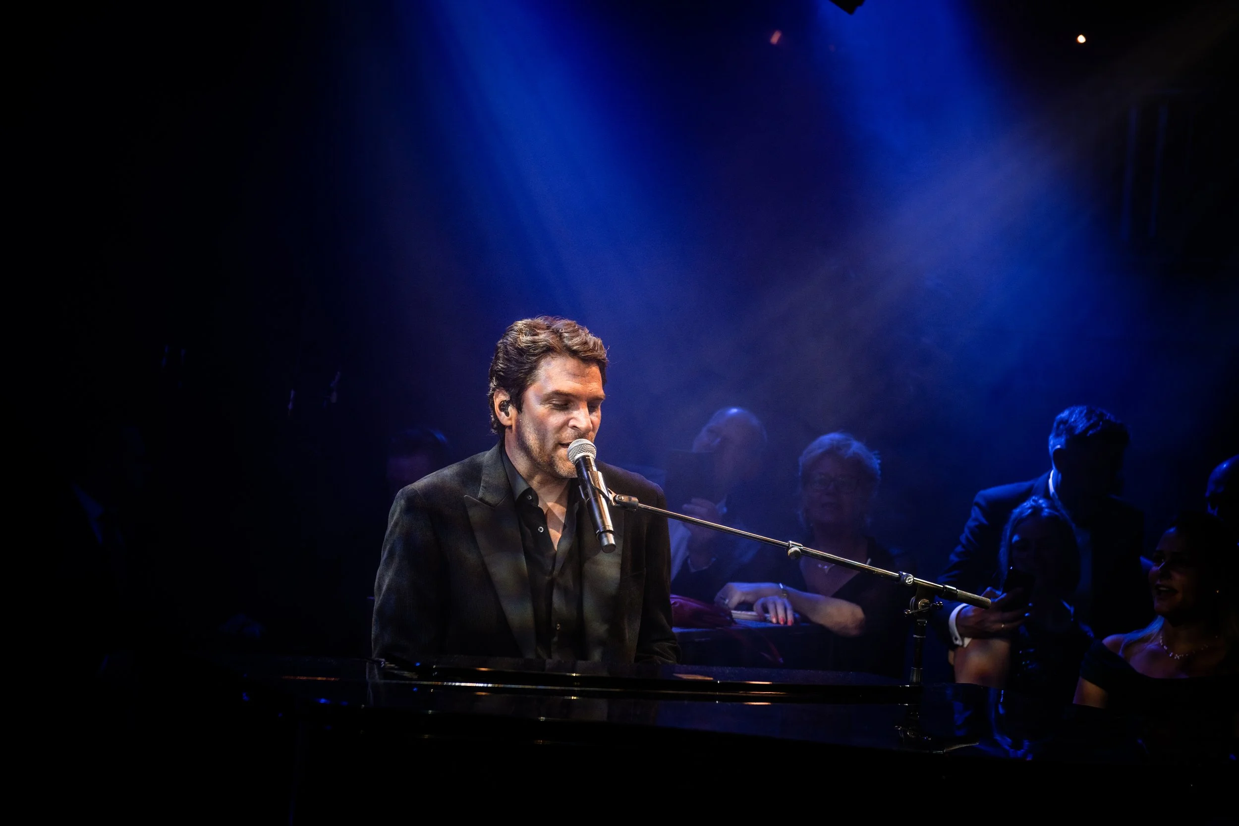 Man die zingt of speelt piano op een podium met blauwe verlichting, omringd door een groep mensen.