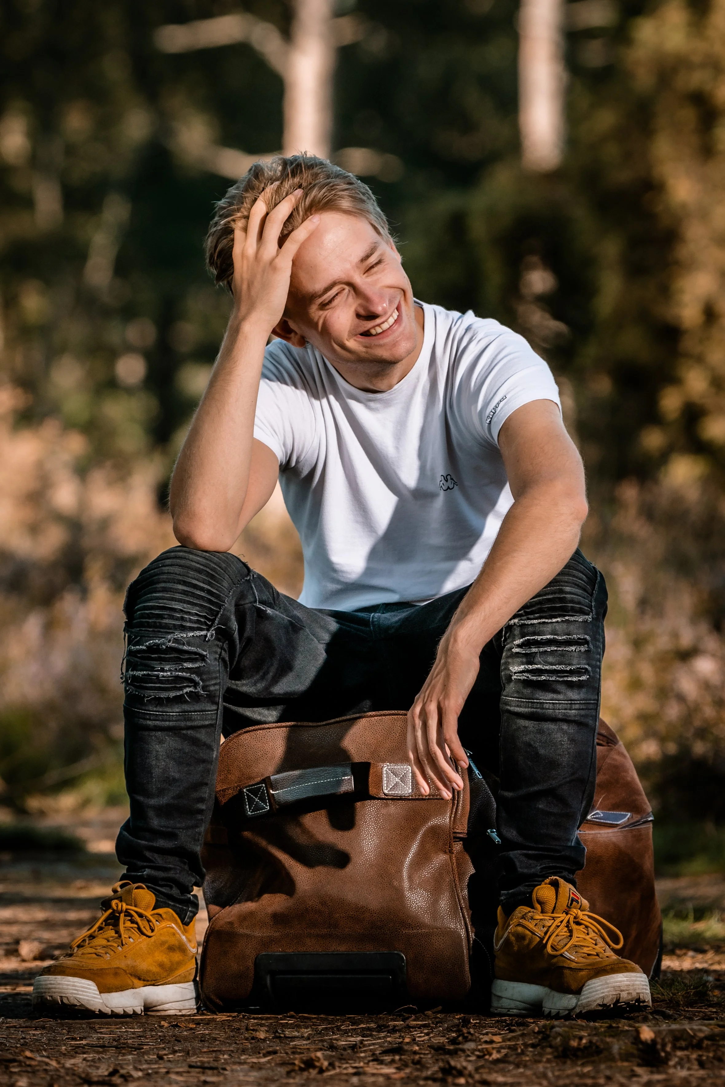 Man lachend, zittend op een reistas in een bos, in een witte T-shirt en zwarte beschadigde spijkerbroek, omgeven door bomen.