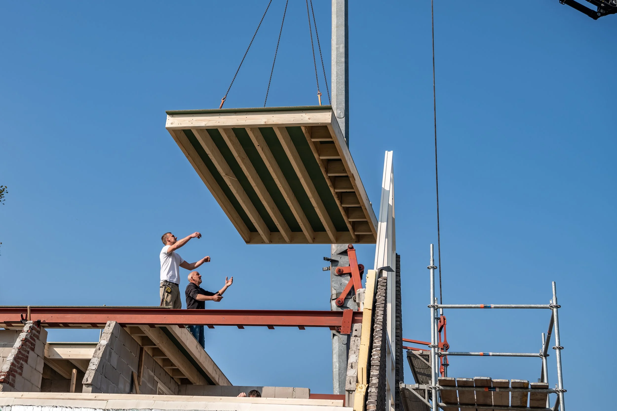 Twee bouwvakkers werken op een dak, één van hen wijst naar het hout en de ander wijst omhoog, onder een heldere blauwe lucht.